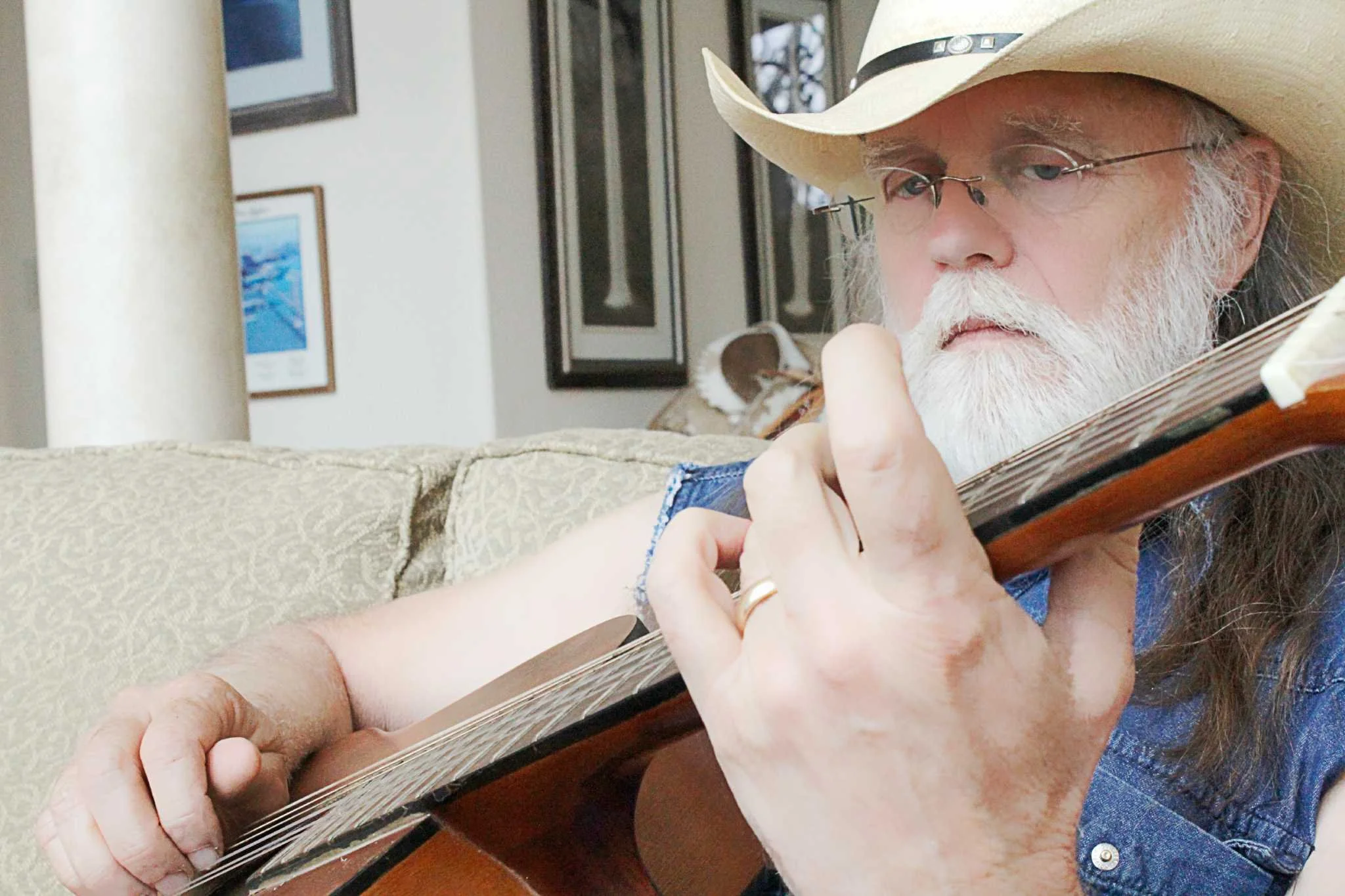 An older man with a white beard, wearing glasses, a cowboy hat, and a denim shirt, playing an acoustic guitar indoors.
