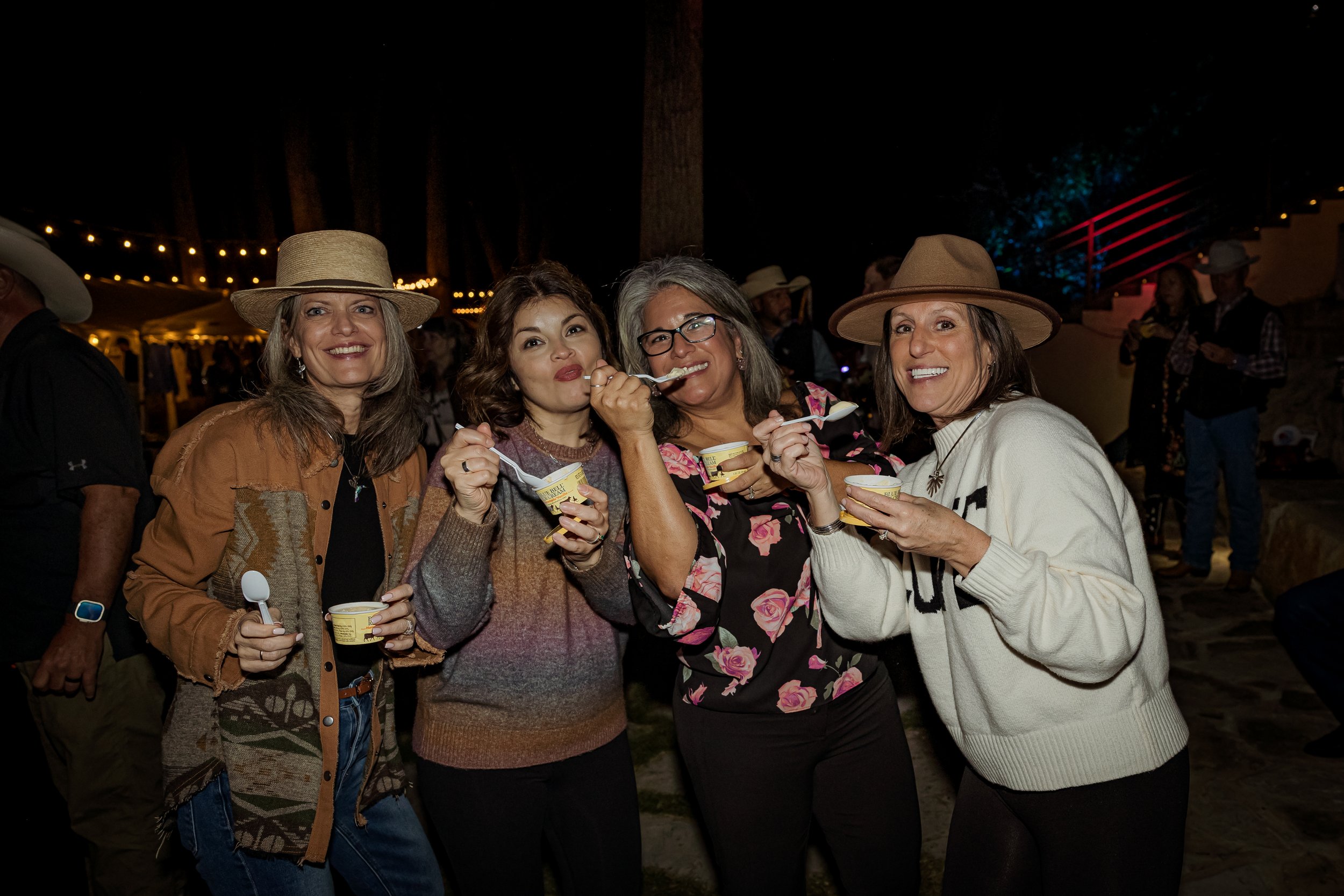 Four women smiling and enjoying ice cream at an outdoor night event, wearing hats and casual clothing, with decorated string lights in the background.