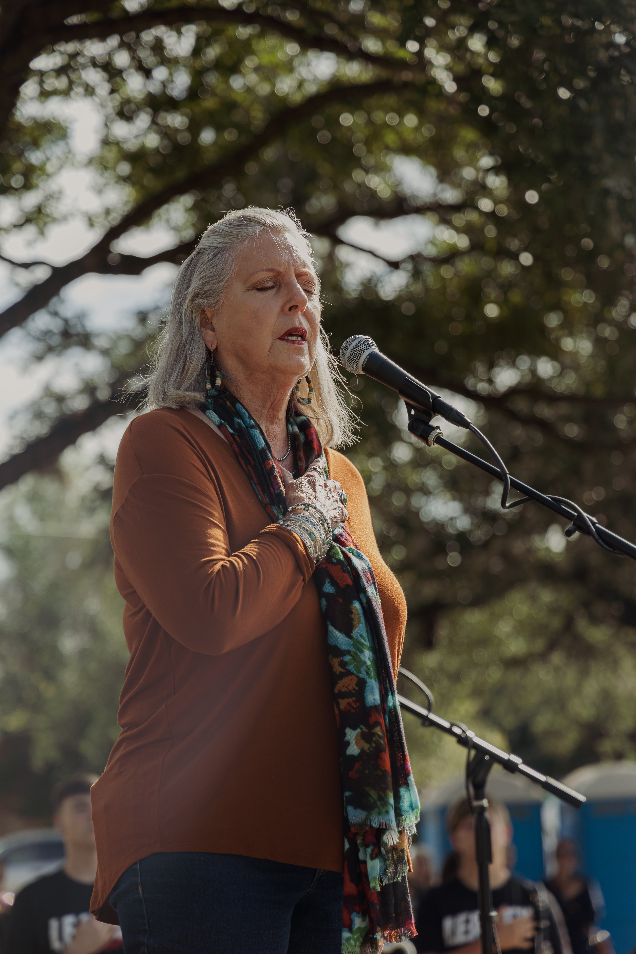 An elderly woman with gray hair singing or speaking into a microphone at an outdoor event, holding her hand over her heart, with trees and people in the background.
