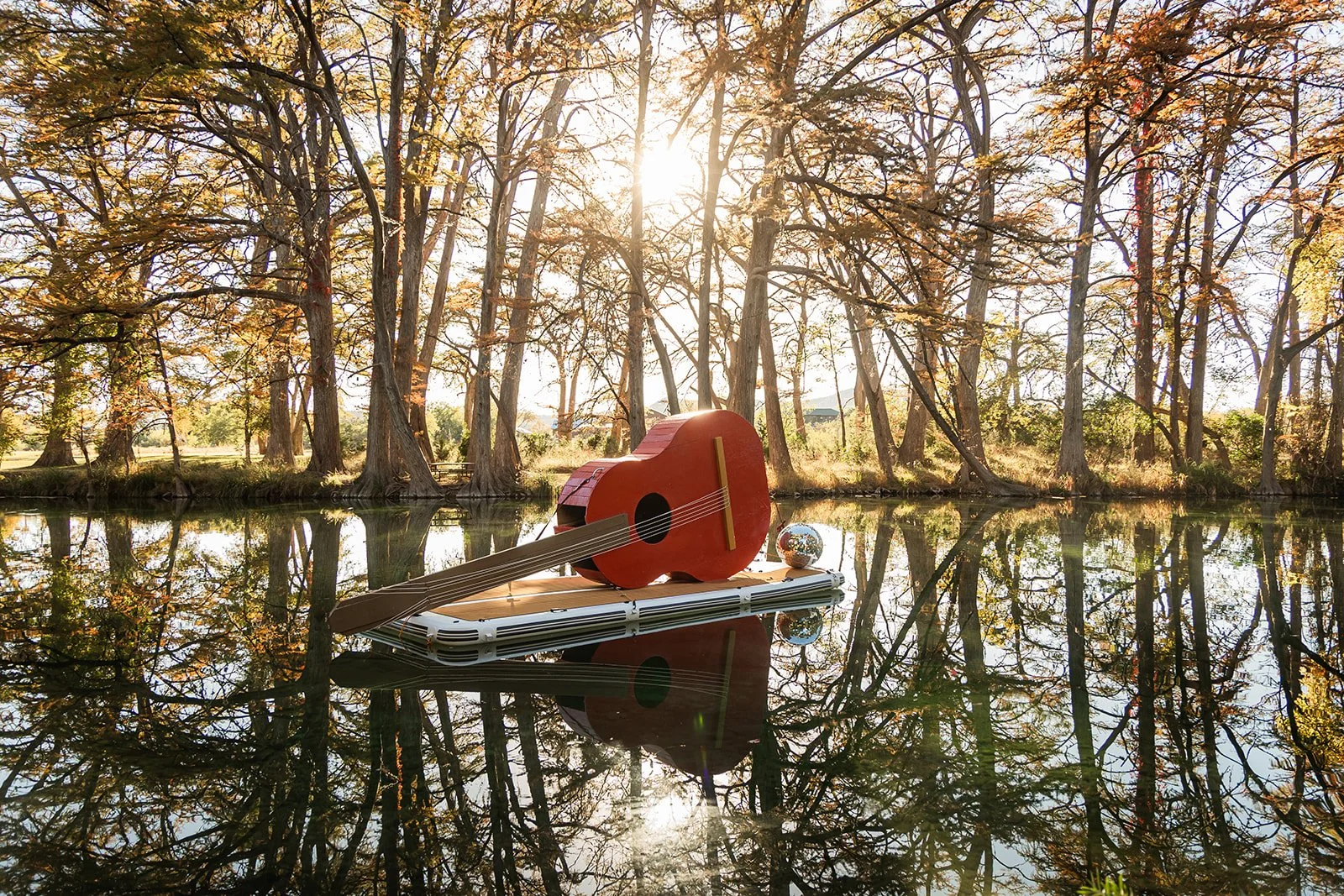 A giant red toy guitar and a small globe sit on a floating platform on a calm lake, with tall trees and sunlight in the background.