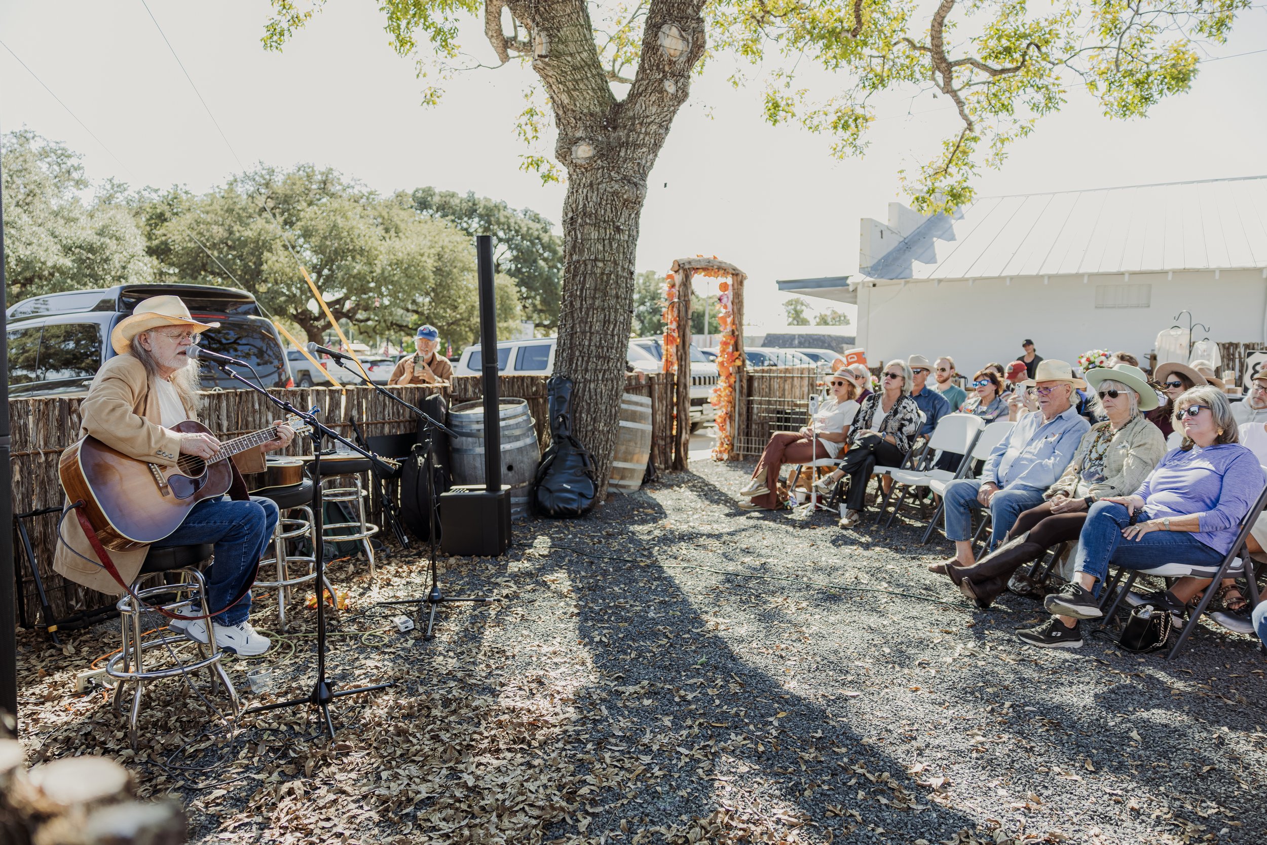 An outdoor concert with an older musician playing guitar and singing into a microphone, seated under a large tree, with an audience sitting on white chairs and some taking photos, set in a rustic, sunny environment.