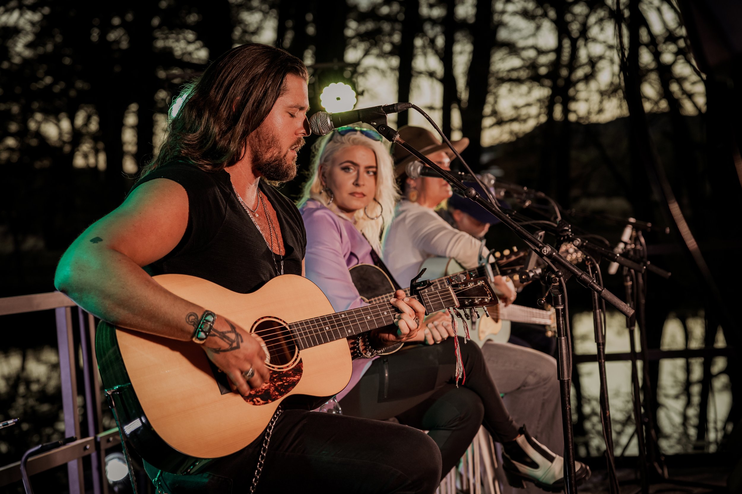 A group of musicians performing outdoors at dusk, with three people playing acoustic guitars and singing into microphones.