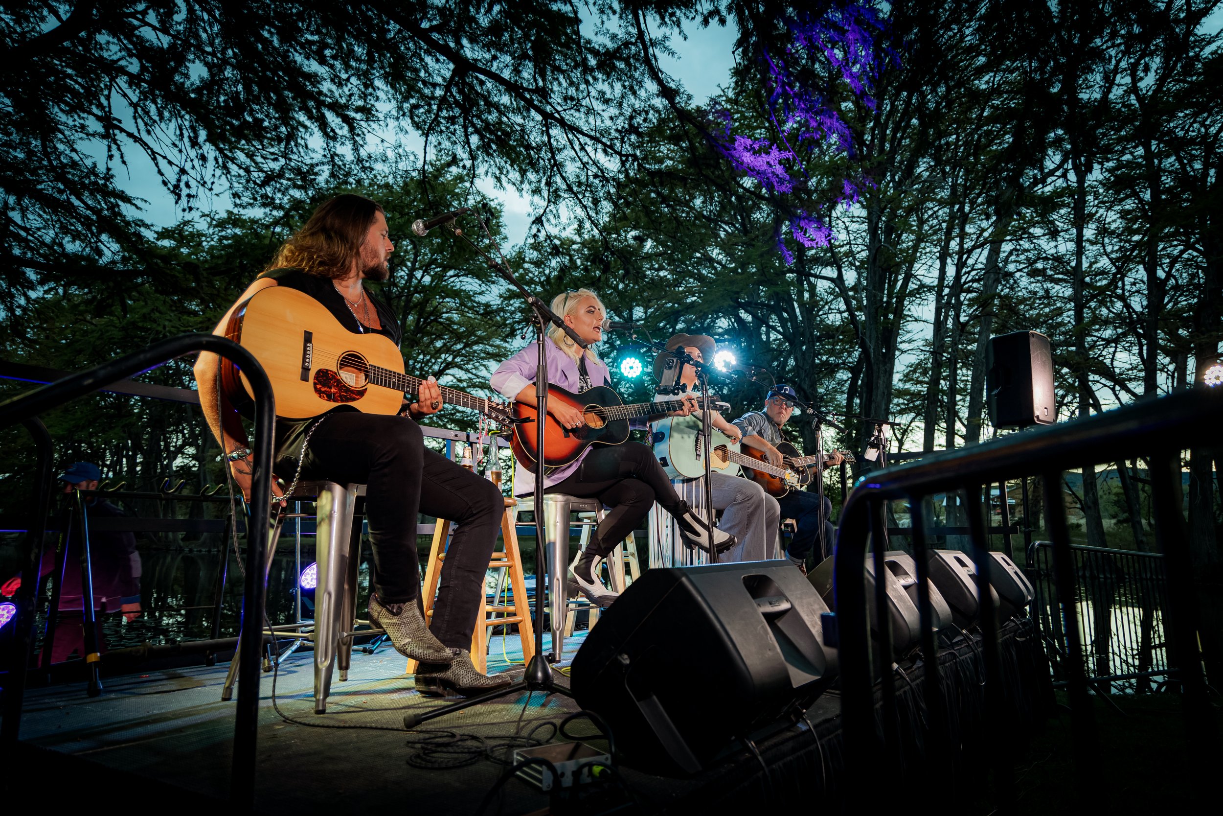 Four musicians seated on a stage playing acoustic guitars during an outdoor concert at dusk, surrounded by trees with purple and blue lighting.