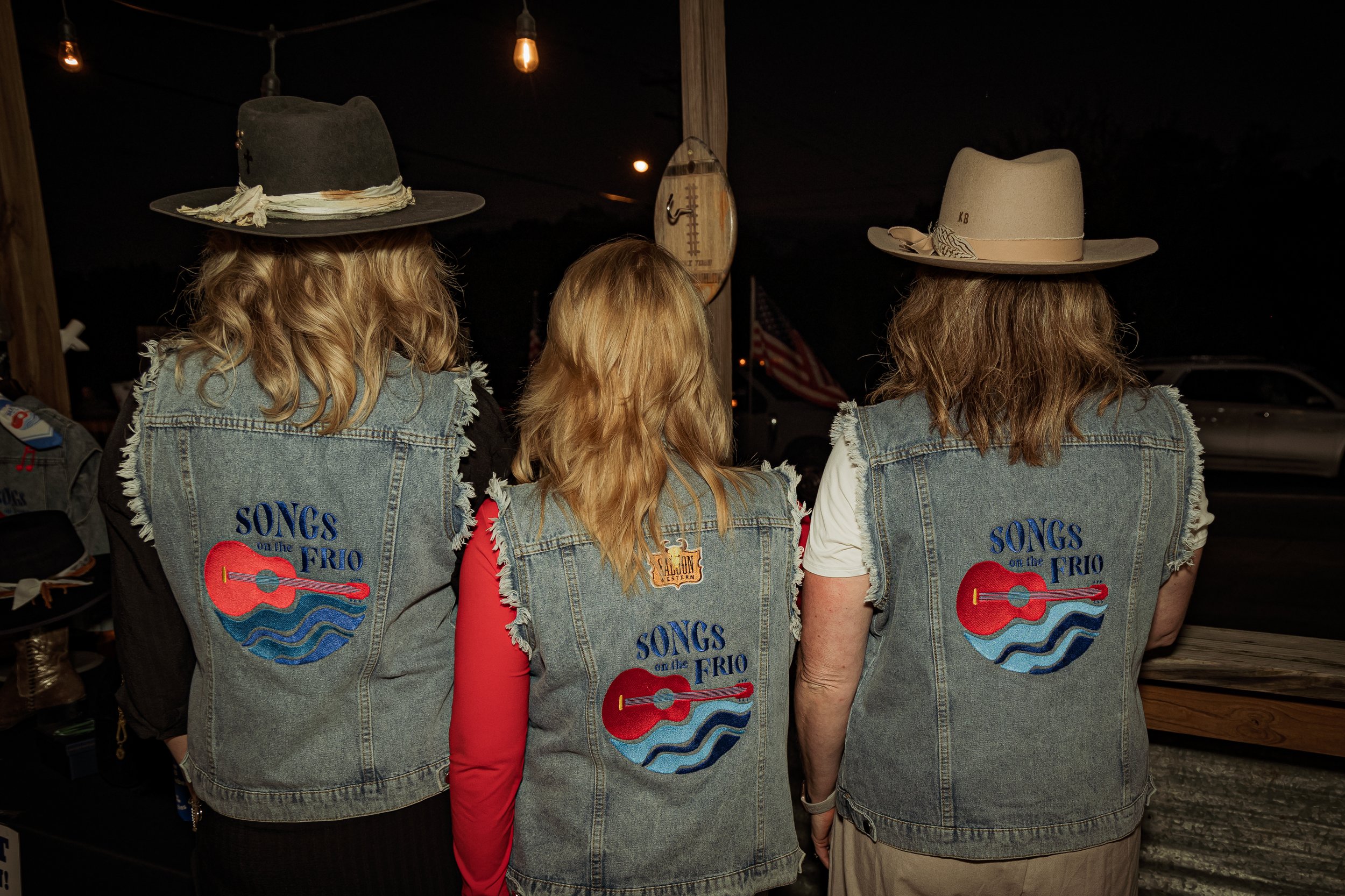 Three women wearing denim vests with a 'Songs on the Frio' logo, two of them wearing cowboy hats, standing with their backs to the camera at night.