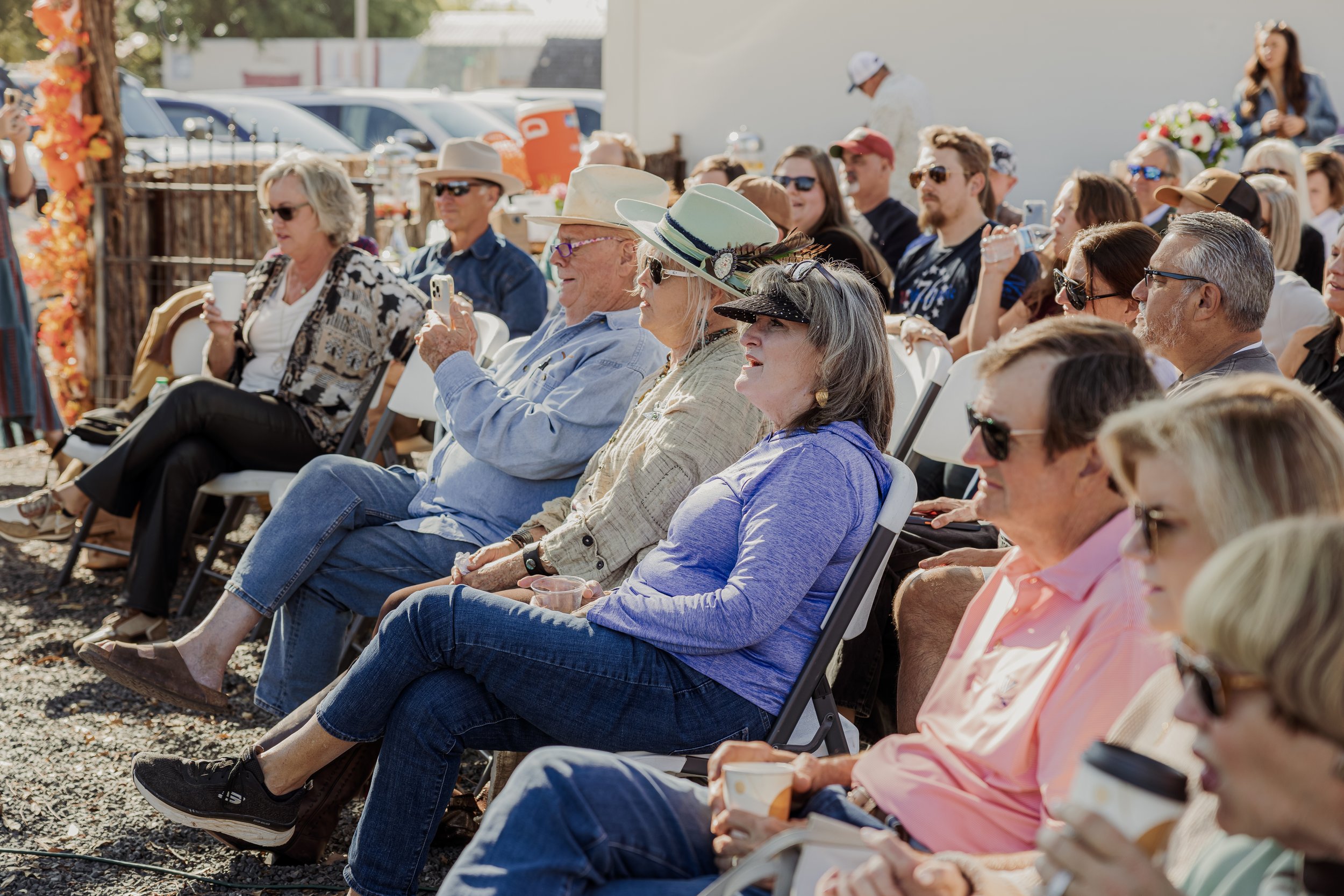 People sitting outdoors at an event, some wearing hats and sunglasses, enjoying drinks, with a backdrop of cars and decorative autumn leaves.