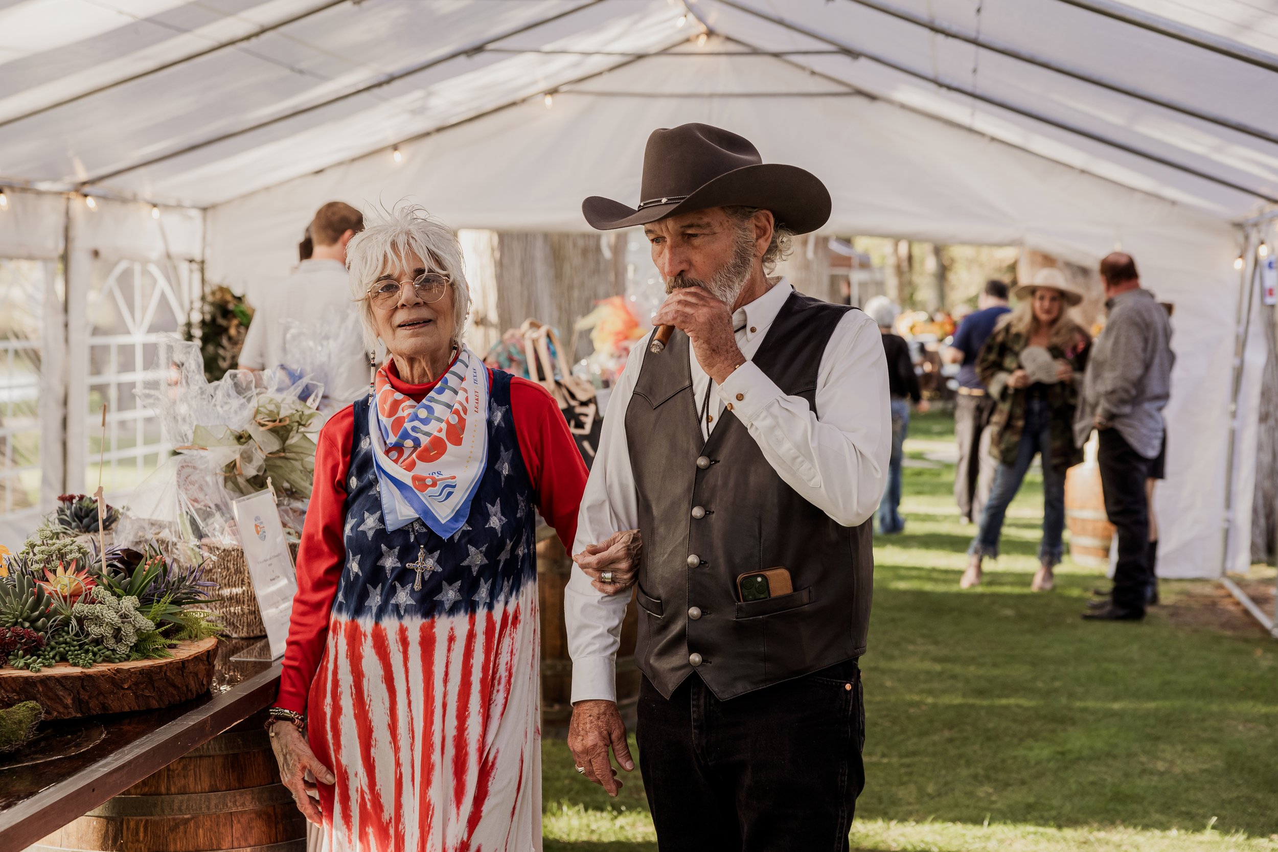 An elderly woman in patriotic American flag dress and a man in cowboy attire smoking a cigar at an outdoor event under a white tent with other people in the background.
