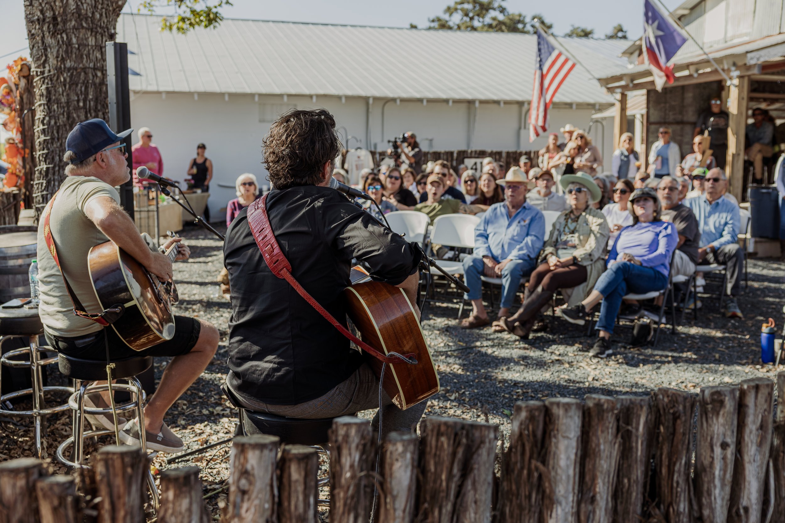 Two musicians performing with guitars in front of seated audience outdoors, some wearing hats and sunglasses, with flags visible in the background.