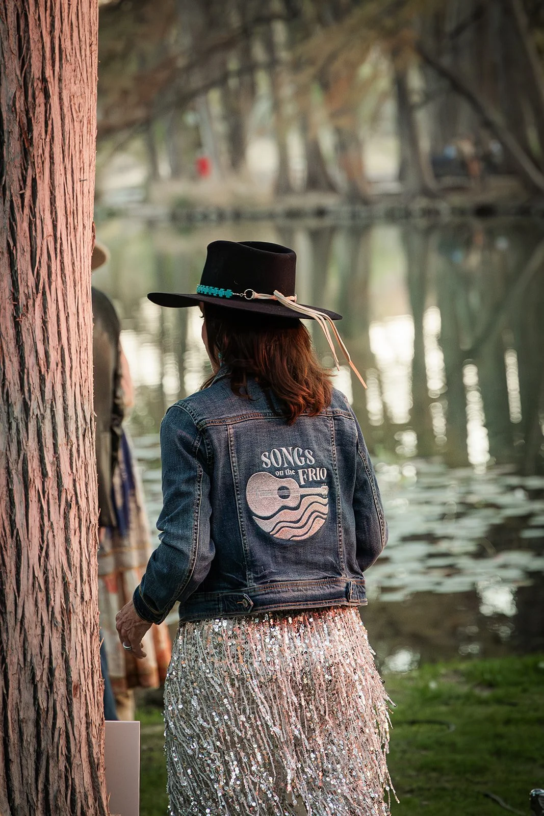 A woman in a denim jacket with a large patch reading 'Songs on the Frio' and a guitar illustration, wearing a black hat with turquoise decorations, standing near a water body with trees and a bridge in the background.