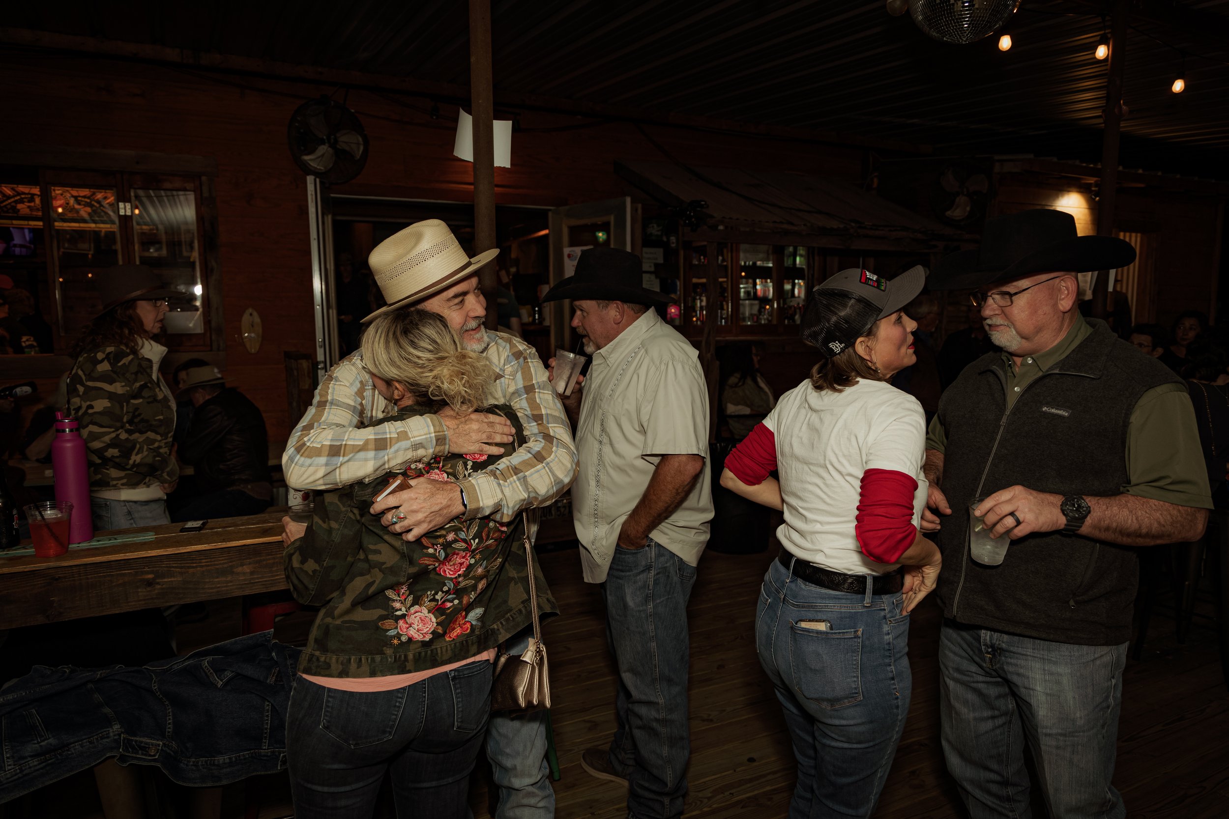 Group of people at a bar, some embracing and chatting, dressed casually with cowboy hats and cowboy-style clothing, in a rustic, dimly-lit setting.