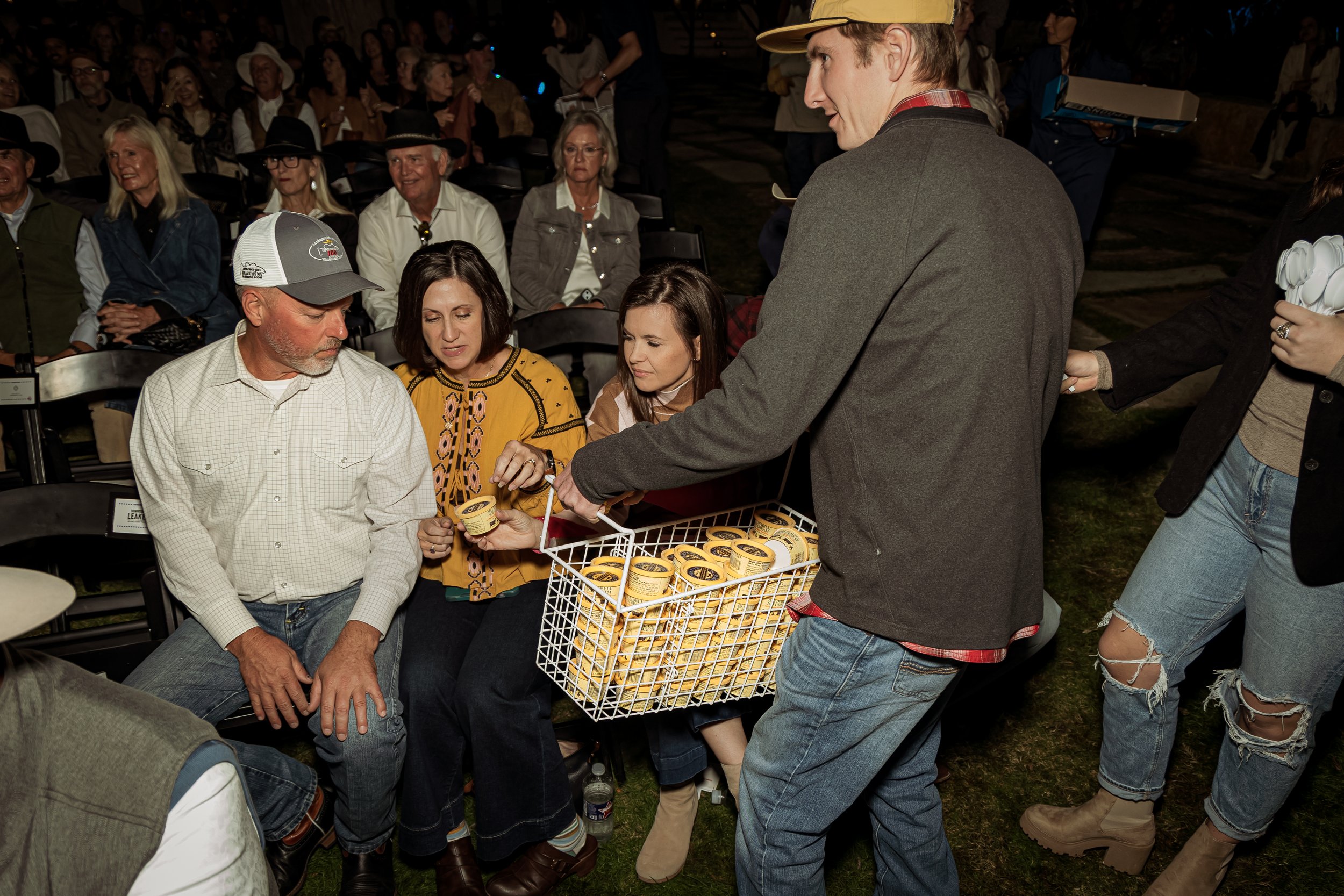People sitting in outdoor chairs at an event, with some receiving yellow containers from a man in a gray jacket.