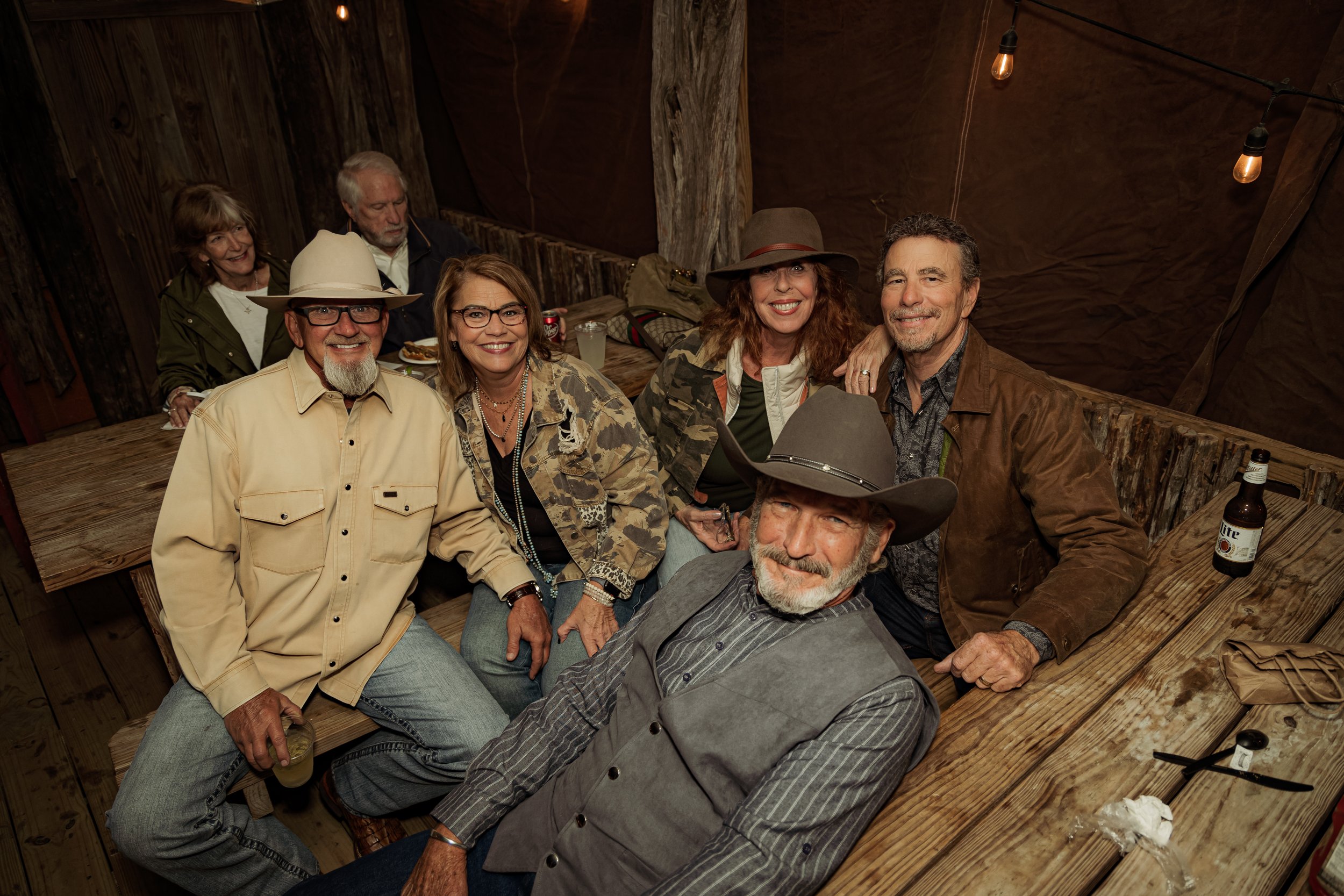 Group of seven people, five adults and two elderly, sitting and standing around a rustic wooden table, smiling in a cozy, dimly lit, rustic setting with string lights.