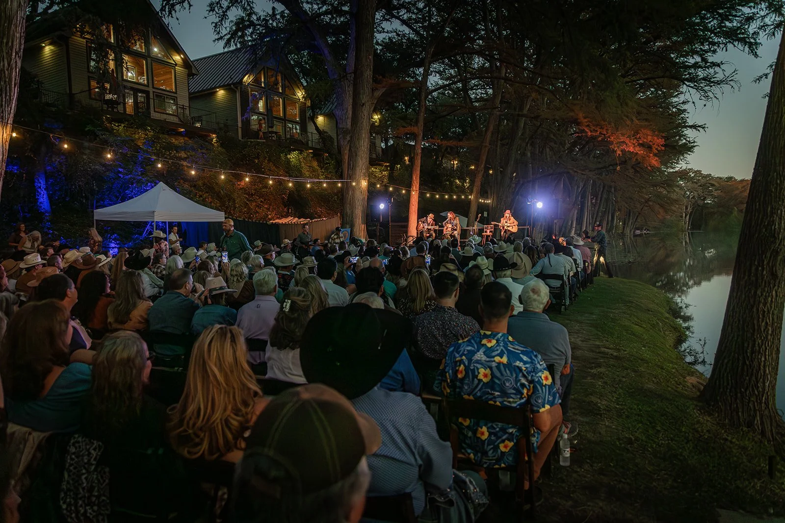 An outdoor concert at dusk with a large audience sitting facing a stage near a river, surrounded by tall trees and illuminated with string lights and stage lights, with a house on the hillside in the background.