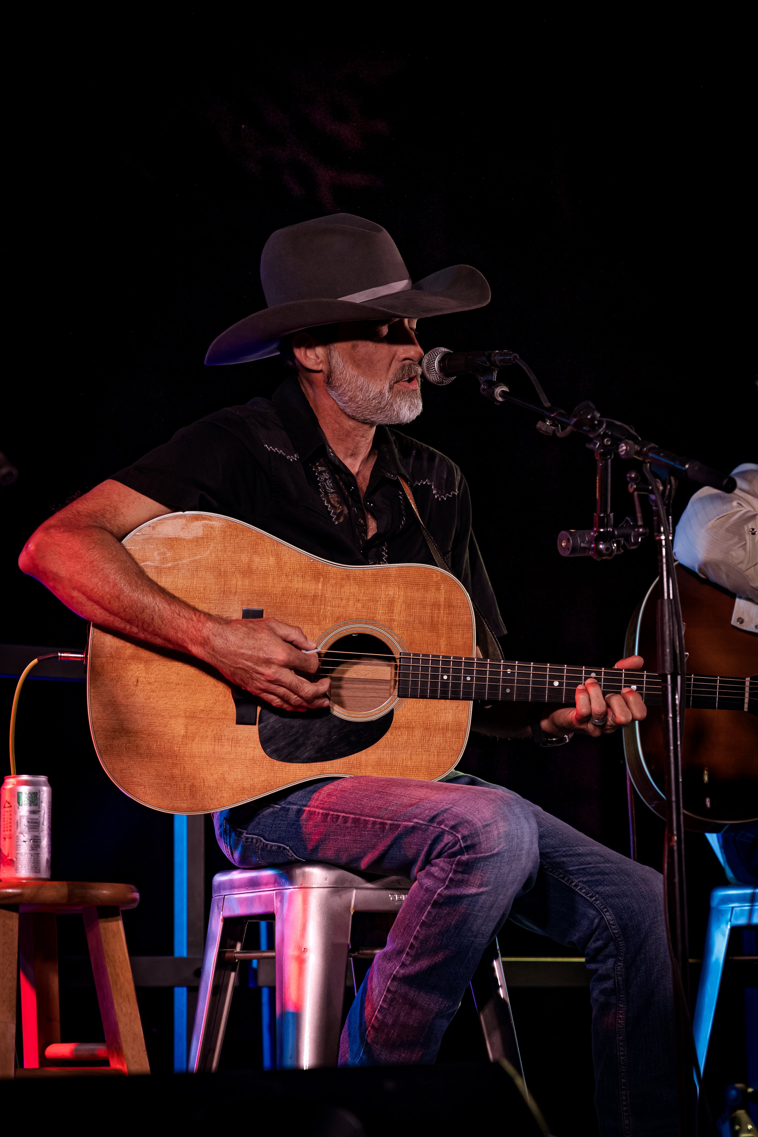 An older man with gray beard and hair, wearing a wide-brimmed hat and sunglasses, plays an acoustic guitar and sings into a microphone during a performance, with black background and colored stage lighting.