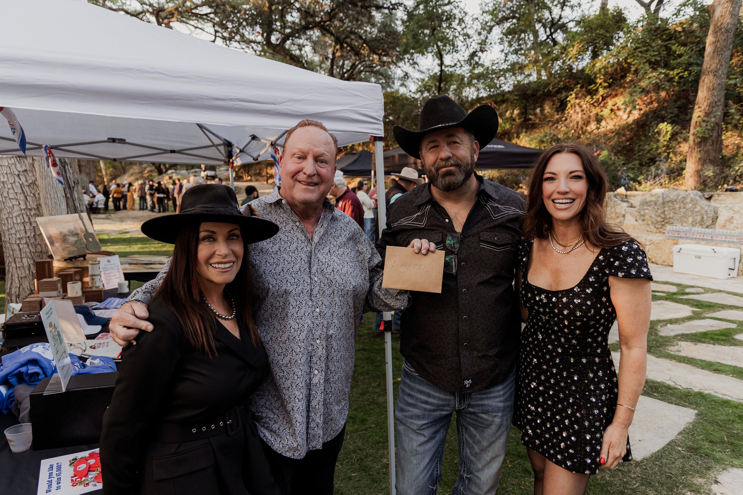 Four people smiling at an outdoor event, two women and two men. One man is holding a paper with '$5000' written on it. Tents and other people are in the background.