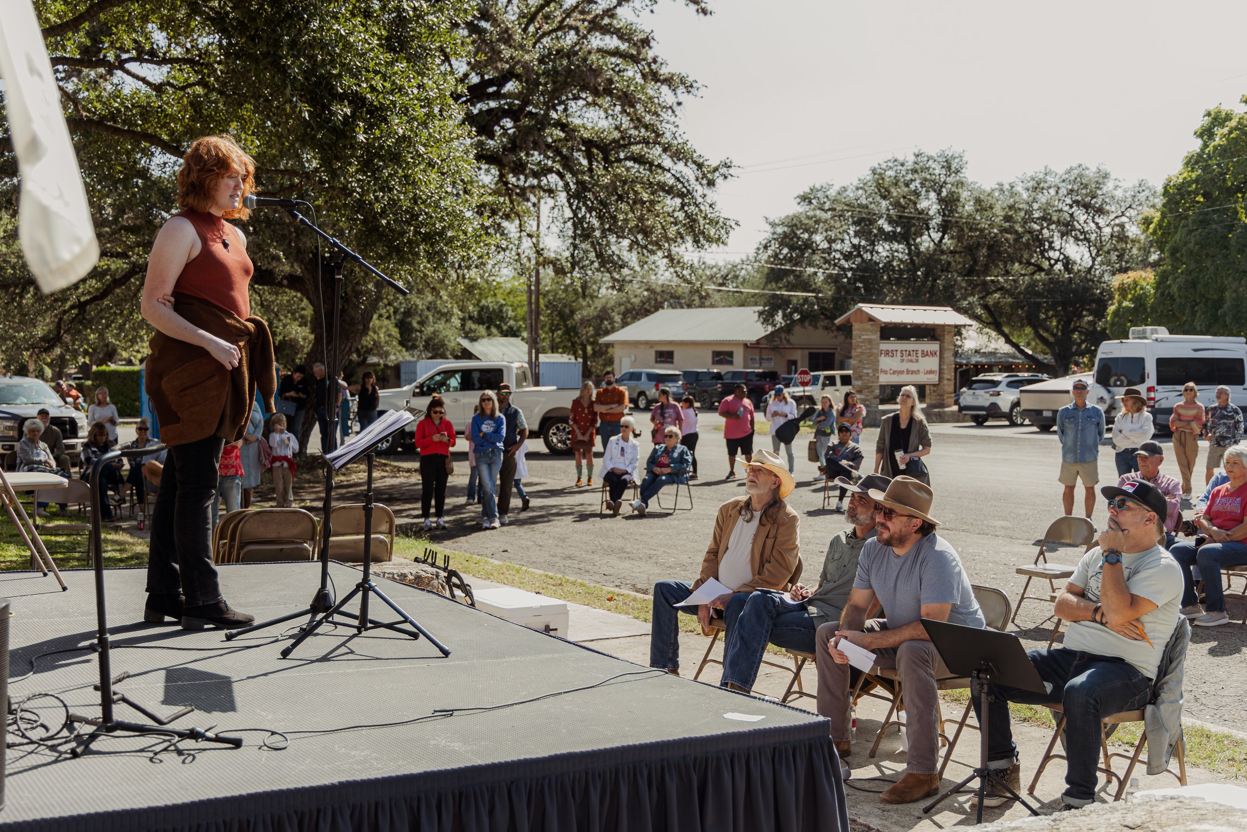 A woman speaking at an outdoor event, with a microphone and music stand, while an audience of mostly older adults sits and stands on a sunny day in a small town.