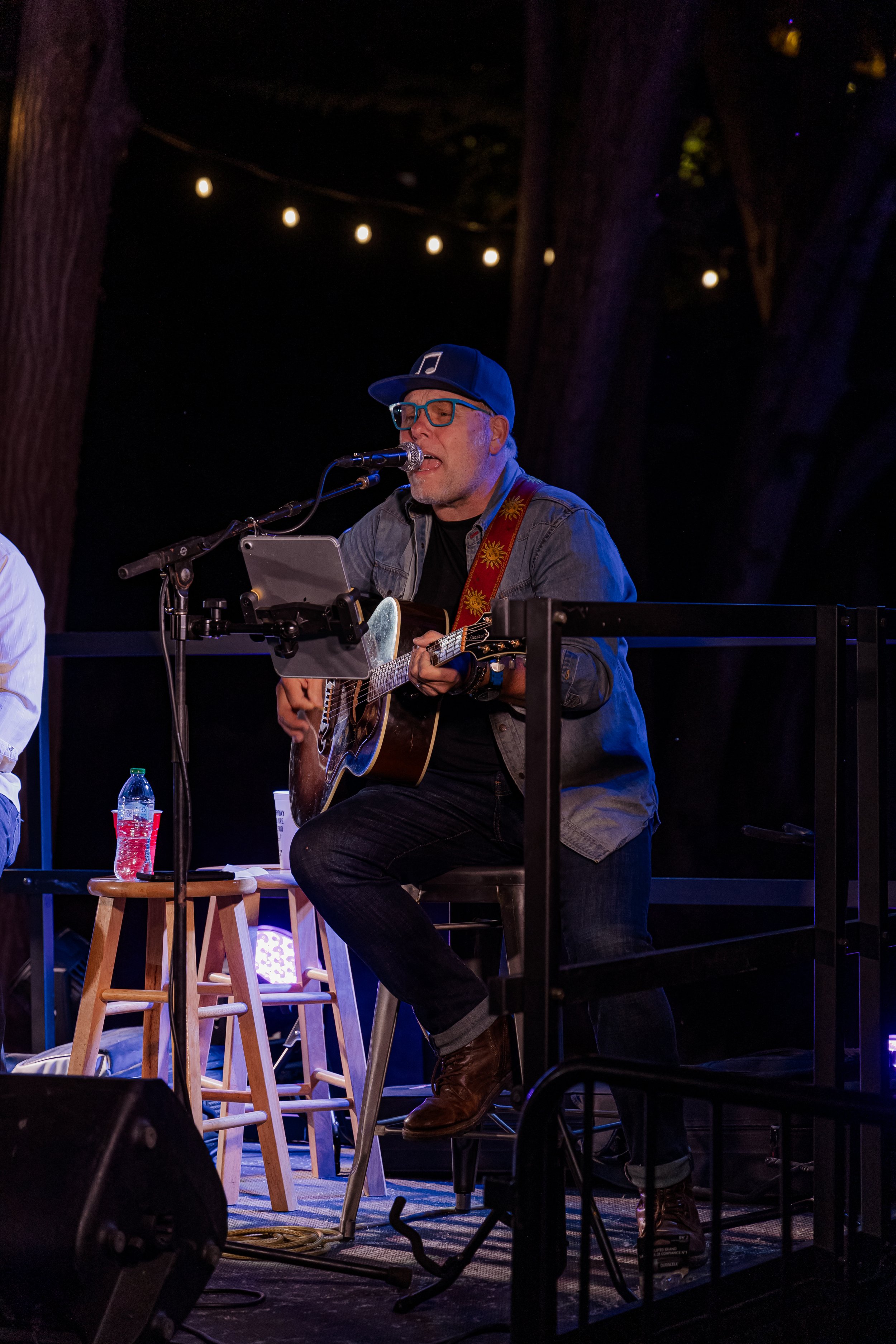 A man wearing glasses, a denim jacket, and a baseball cap is performing on stage, playing an acoustic guitar and singing into a microphone, with string lights overhead.