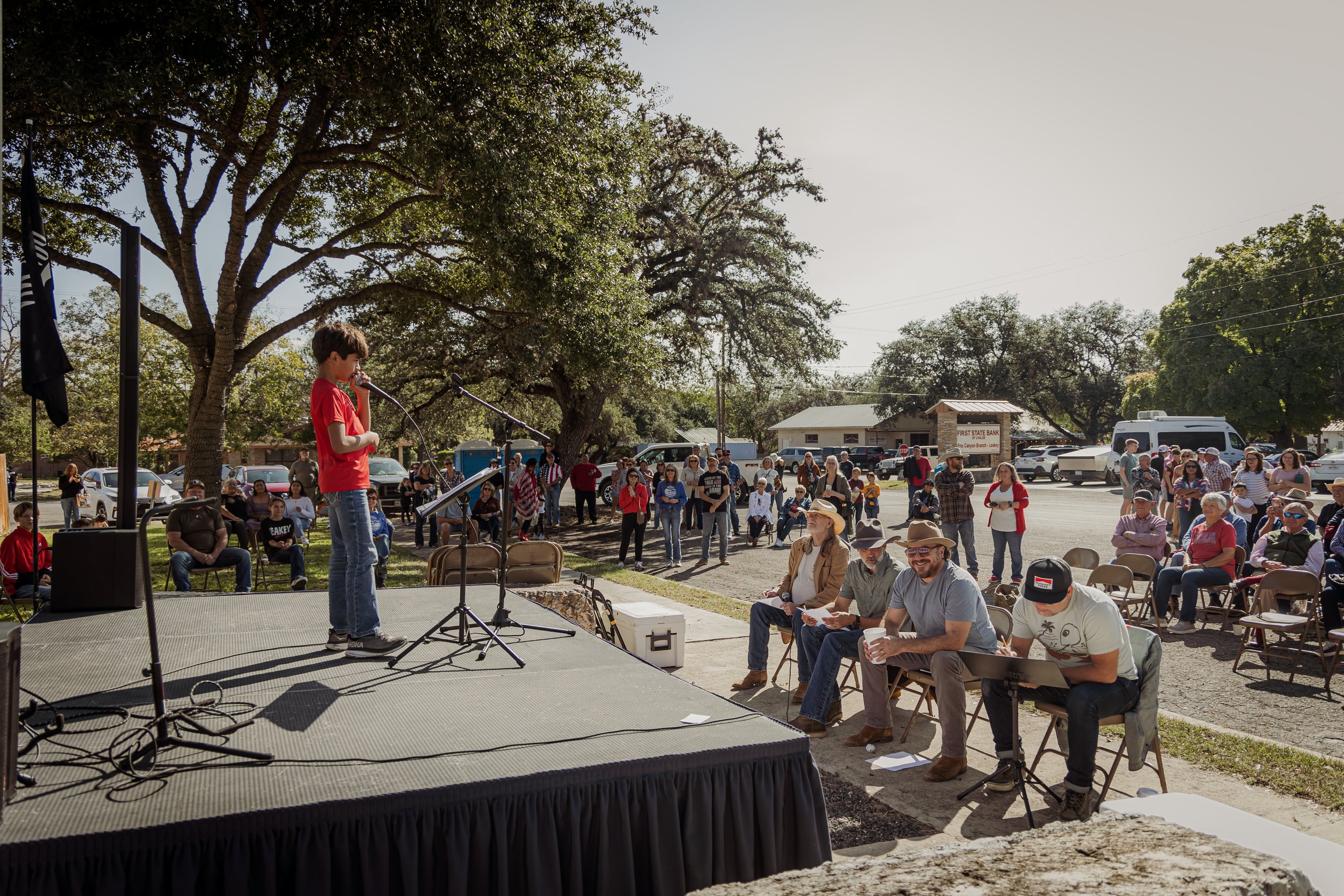 A young boy in a red shirt and jeans speaking into a microphone on a stage at a community outdoor event, with an audience of adults and children seated and standing under trees in the background.