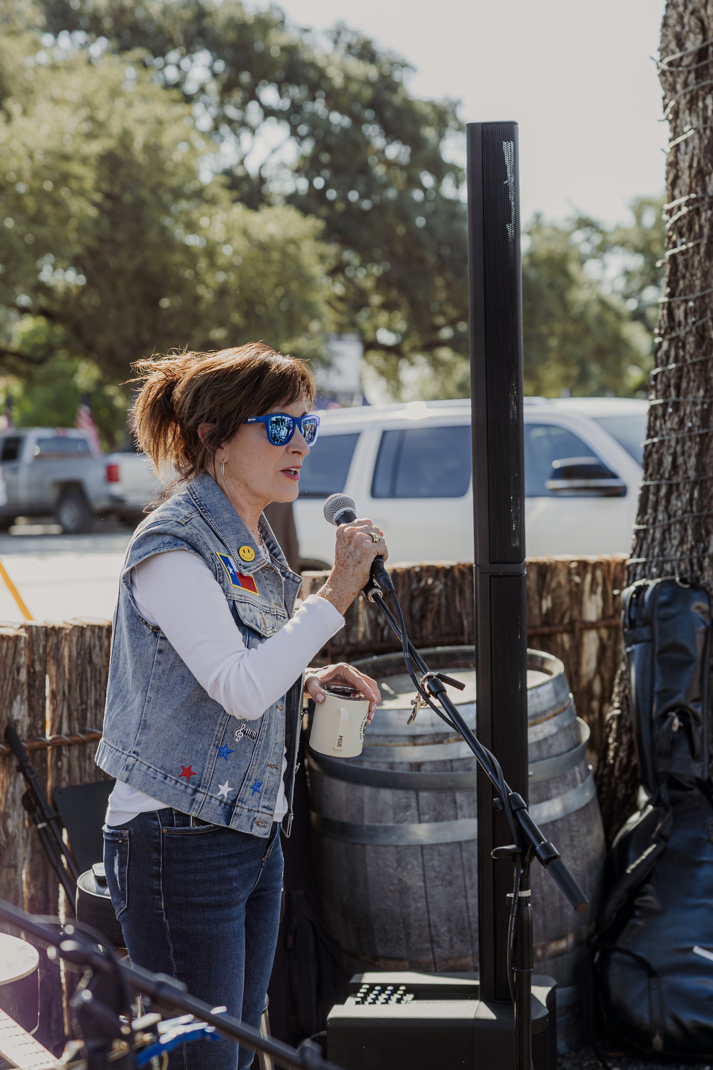 A woman in sunglasses holds a microphone and speaks at an outdoor event with a large speaker, wooden barrels, and parked cars in the background.