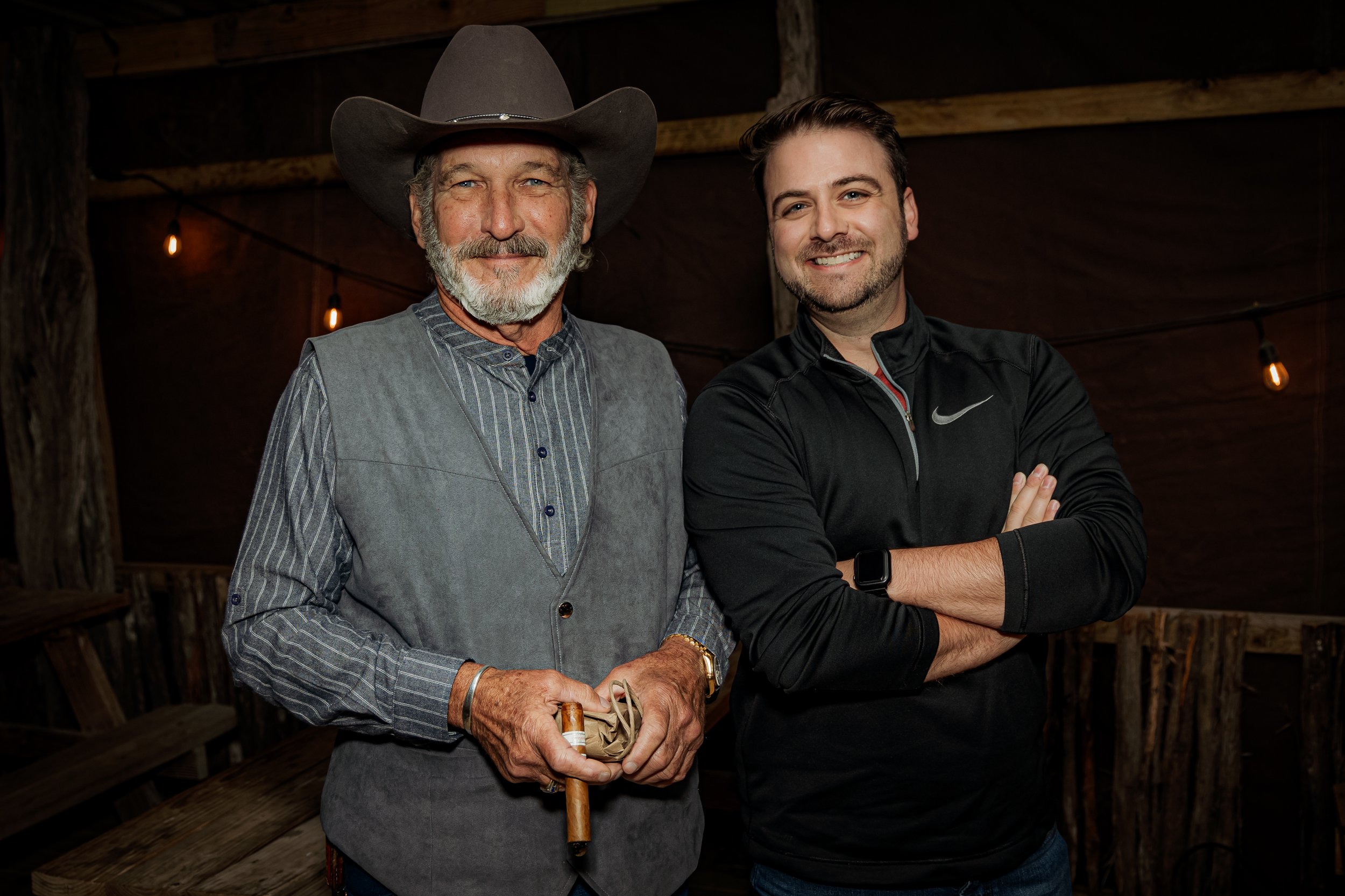 Two men standing together indoors, smiling at the camera. The older man on the left is wearing a cowboy hat, a striped shirt, a vest, and is holding a cigar. The younger man on the right is wearing a black Nike jacket with arms crossed. The backgroun