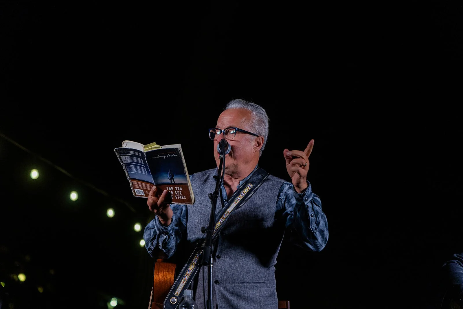 An older man with glasses and grey hair, wearing a grey vest over a blue shirt, is reading from a book into a microphone while holding a guitar. The background is dark with some string lights visible on the left.