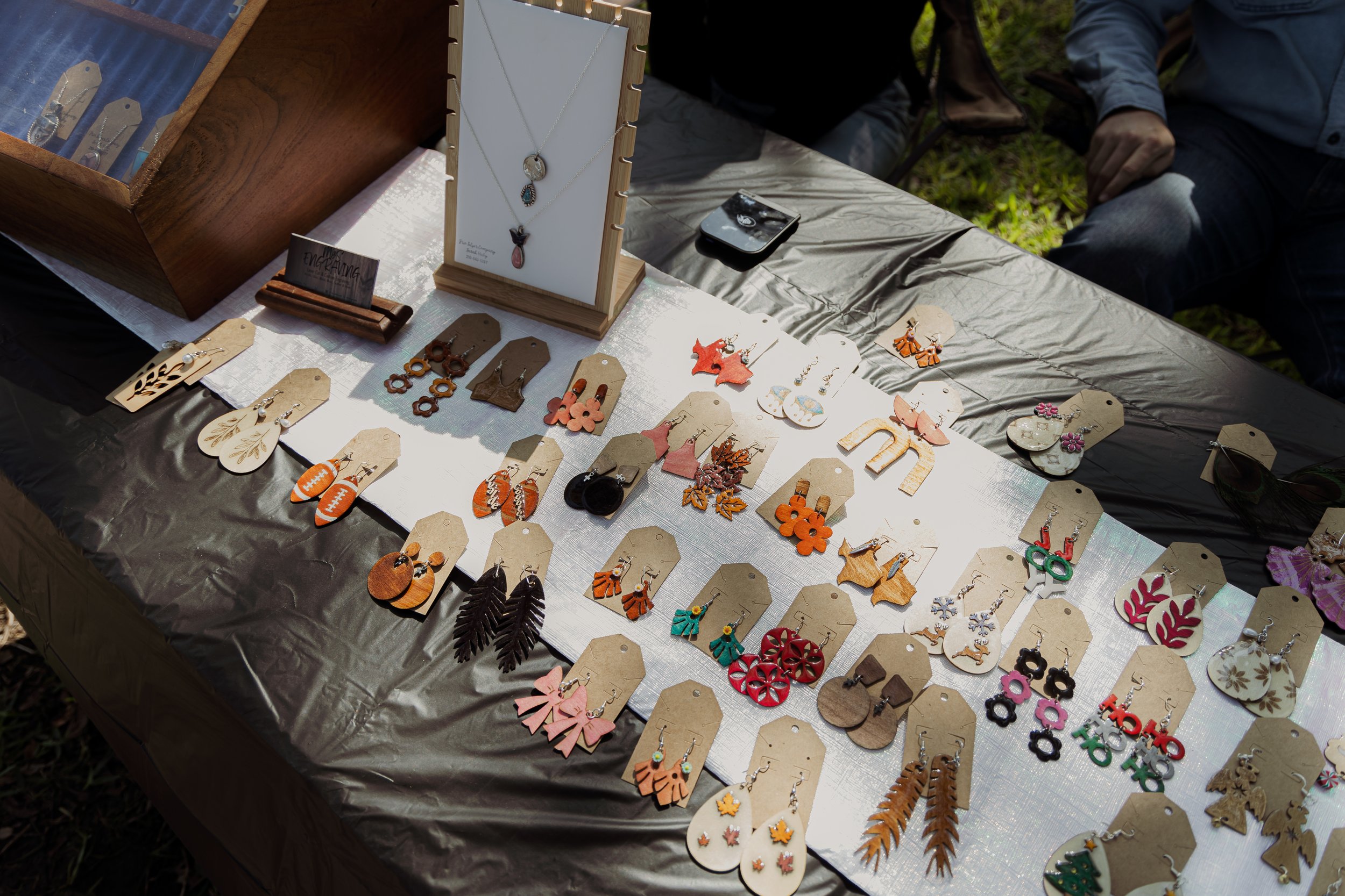 A table at an outdoor craft market displaying various handmade earrings and necklaces on beige display cards, with a wooden jewelry display stand and a black smartphone nearby.
