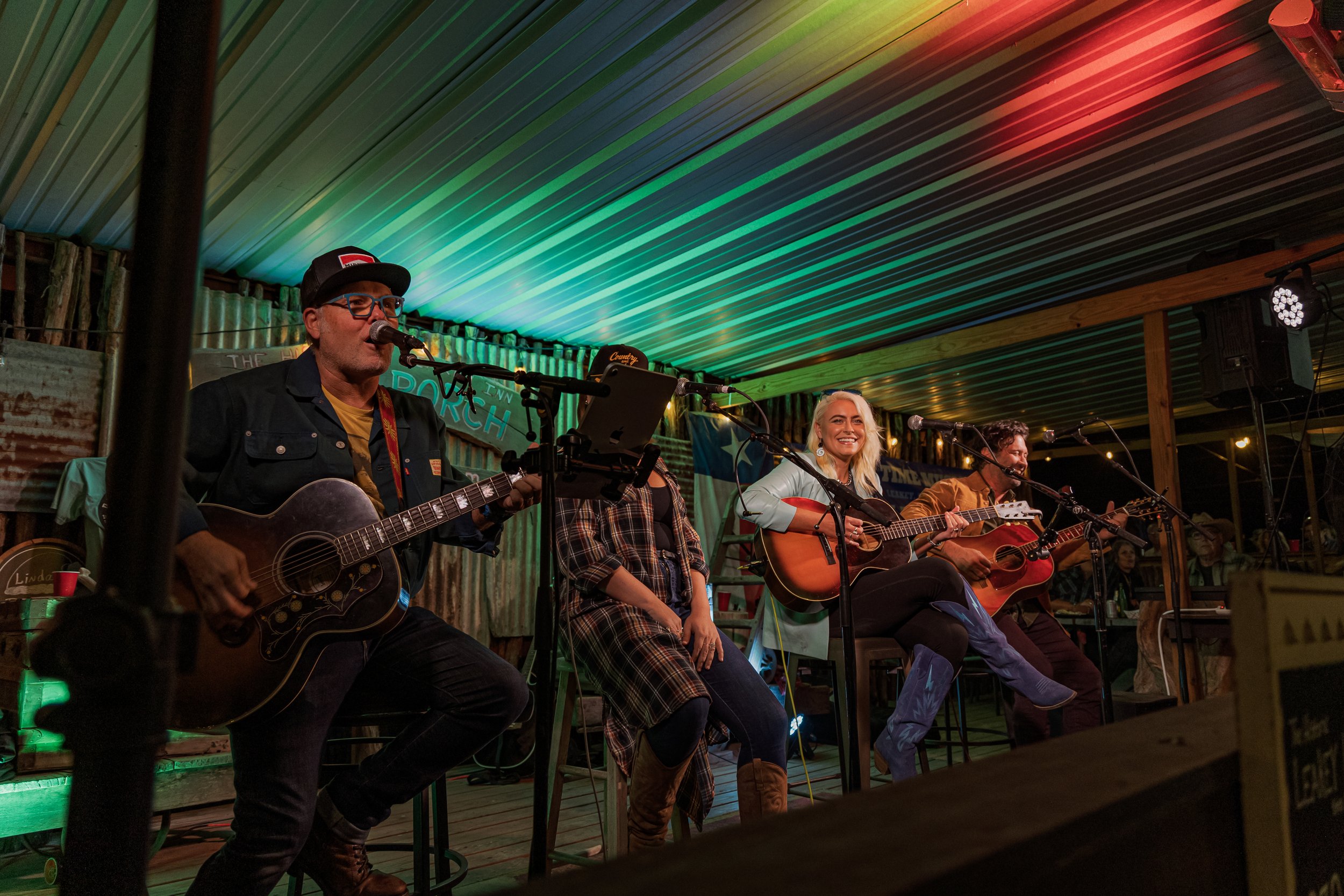 A group of musicians performing on stage with guitars, women and men, in a rustic indoor venue with colorful lighting and audience members visible in the background.