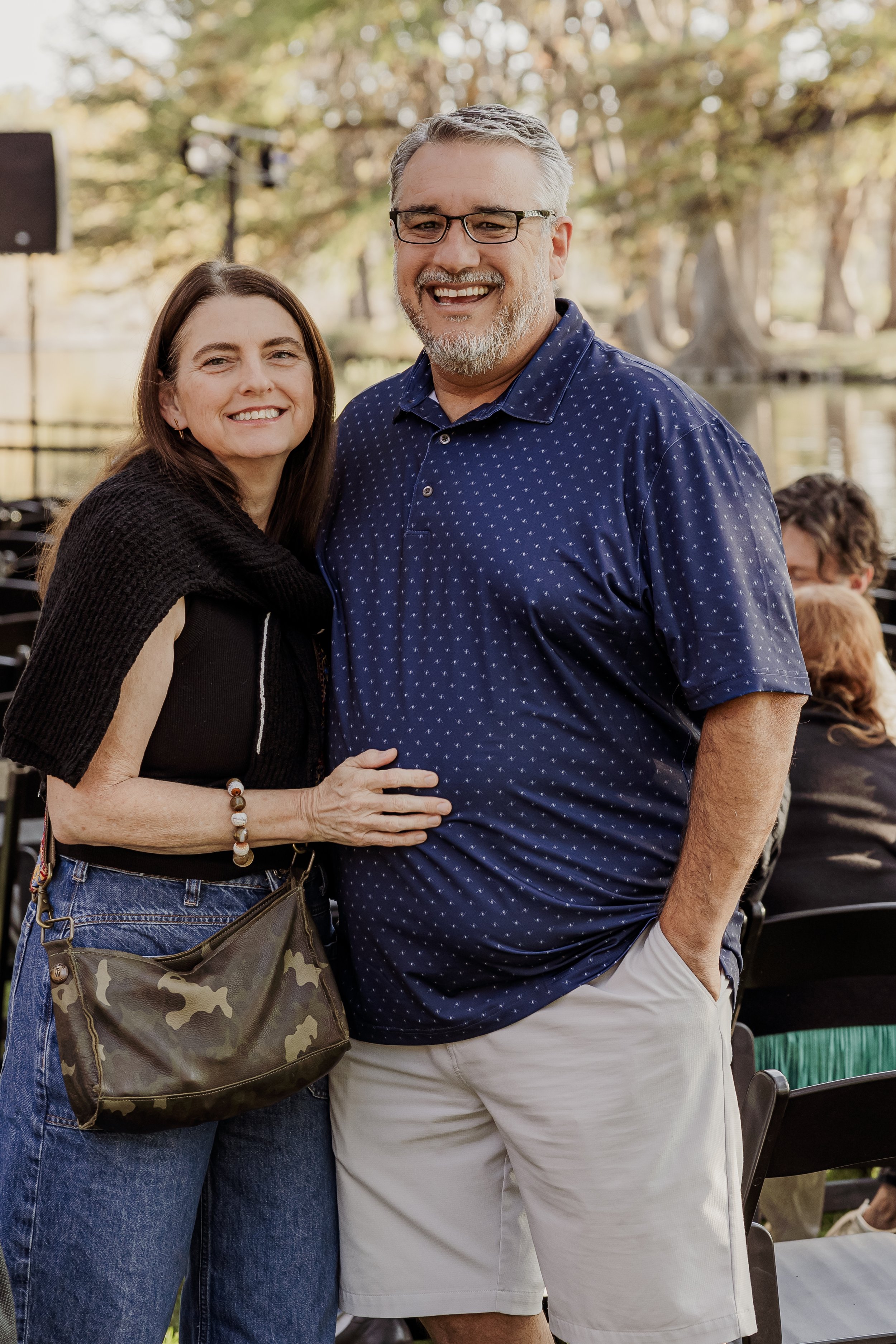 A smiling woman and man standing outdoors near a lake, with trees in the background.
