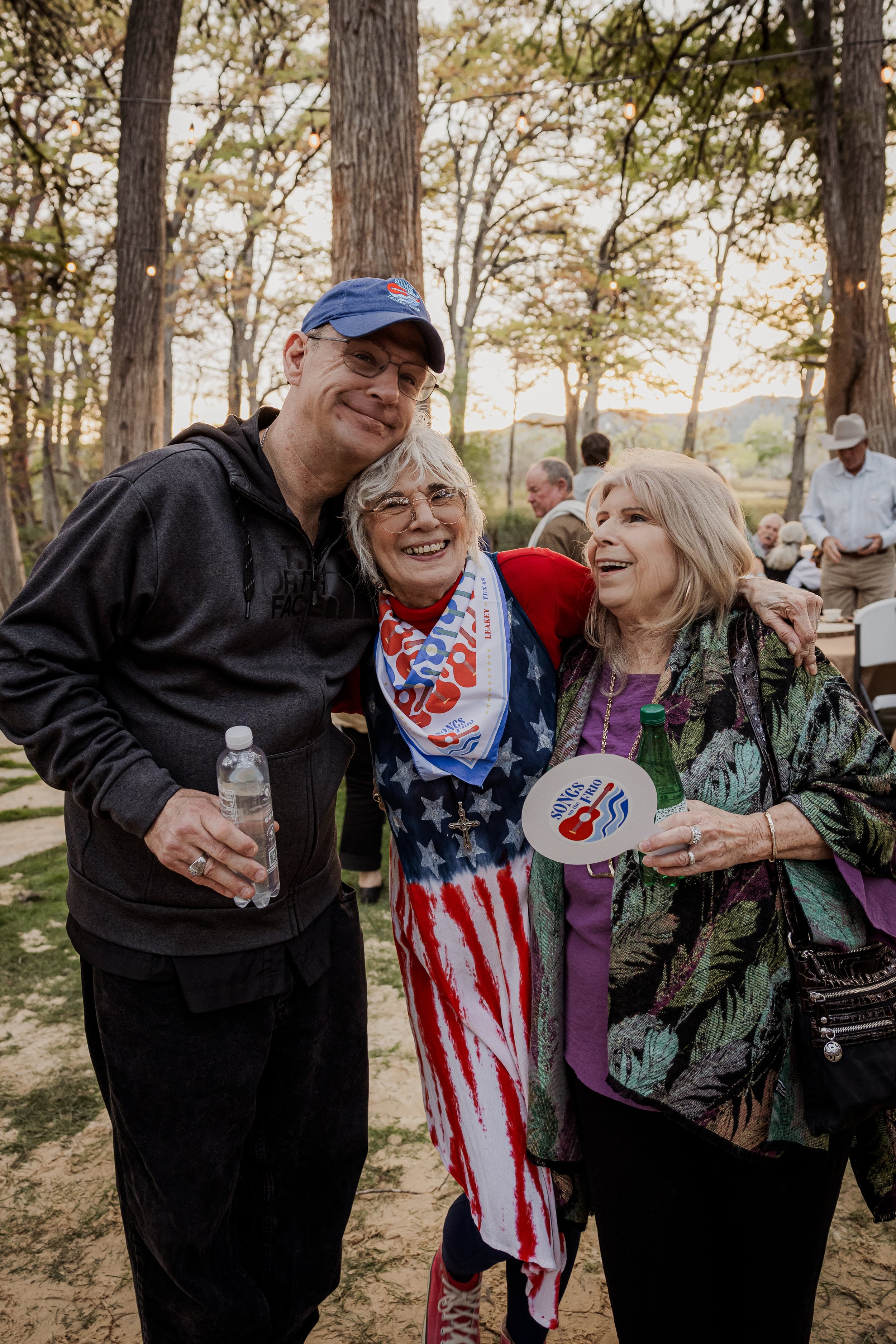 Three people smiling and hugging at an outdoor event, with trees and string lights in the background. The woman in the middle is wearing a patriotic dress and a scarf, holding a fan, and the woman on the right is holding a beer bottle and a fan. The