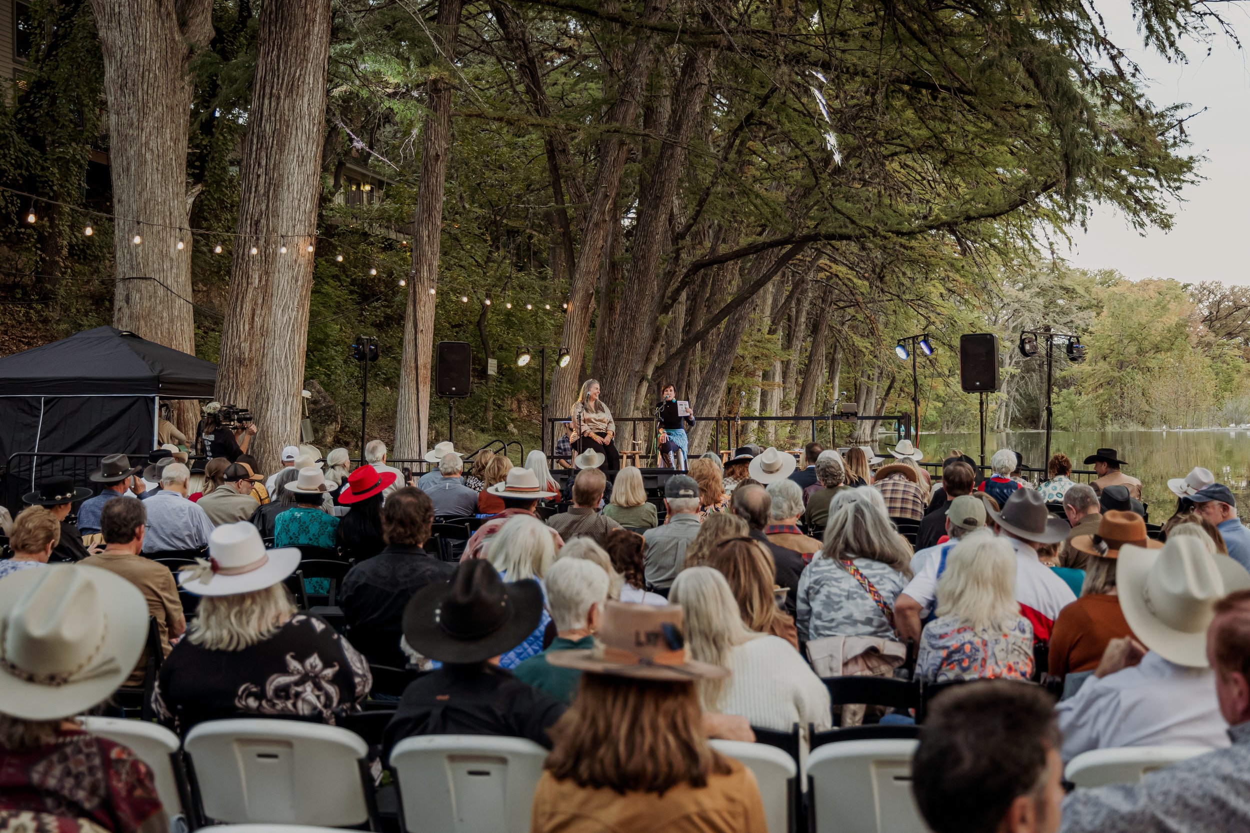 Outdoor audience gathered by a lake listening to a live performance on a small stage surrounded by large trees and string lights.