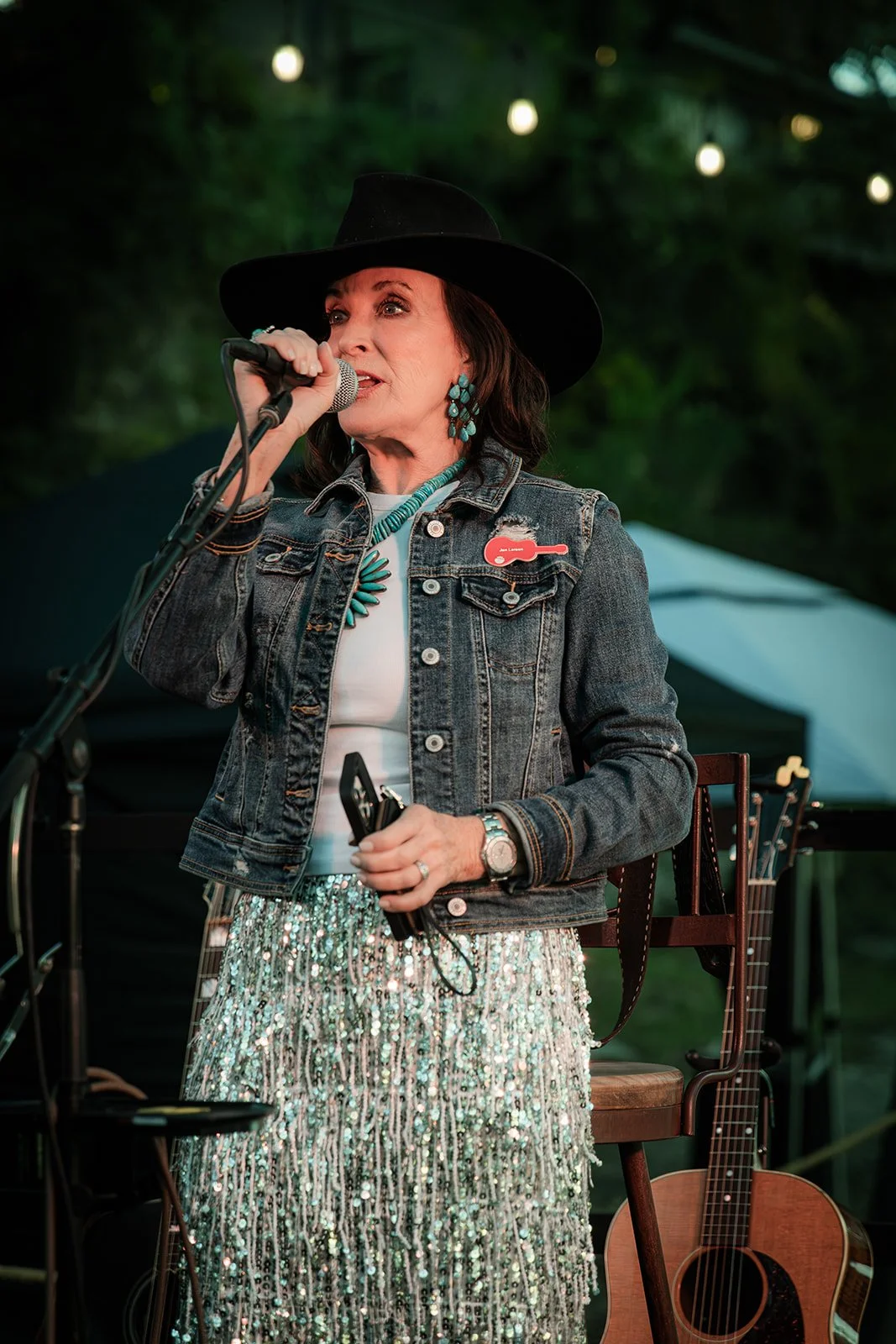Woman in black hat and denim jacket singing into microphone at outdoor event, with guitar and tent in background.