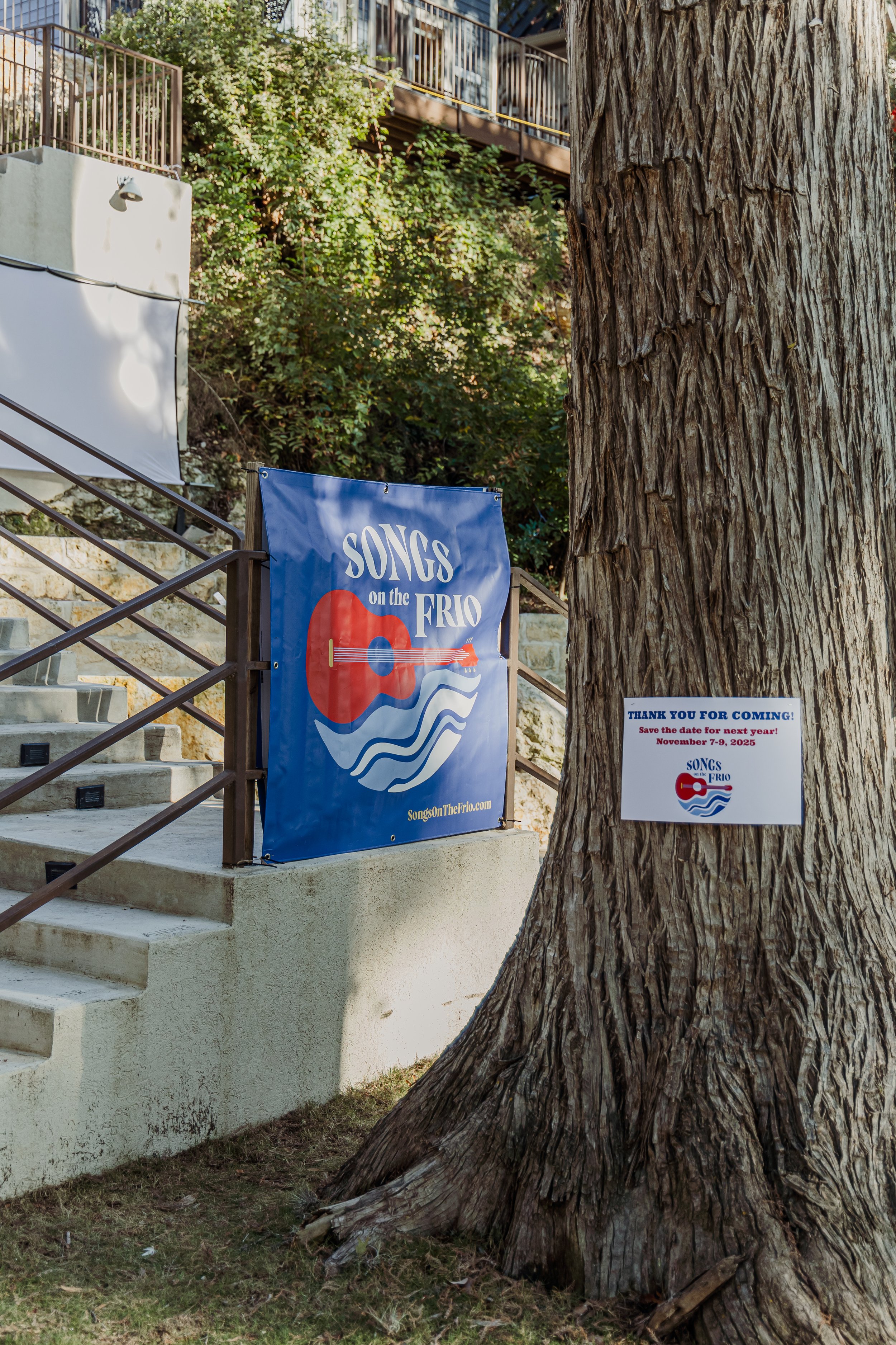 A sign for an event called 'Songs on the Frio' hangs near a large tree, with another larger banner nearby, indicating the event's website and date.
