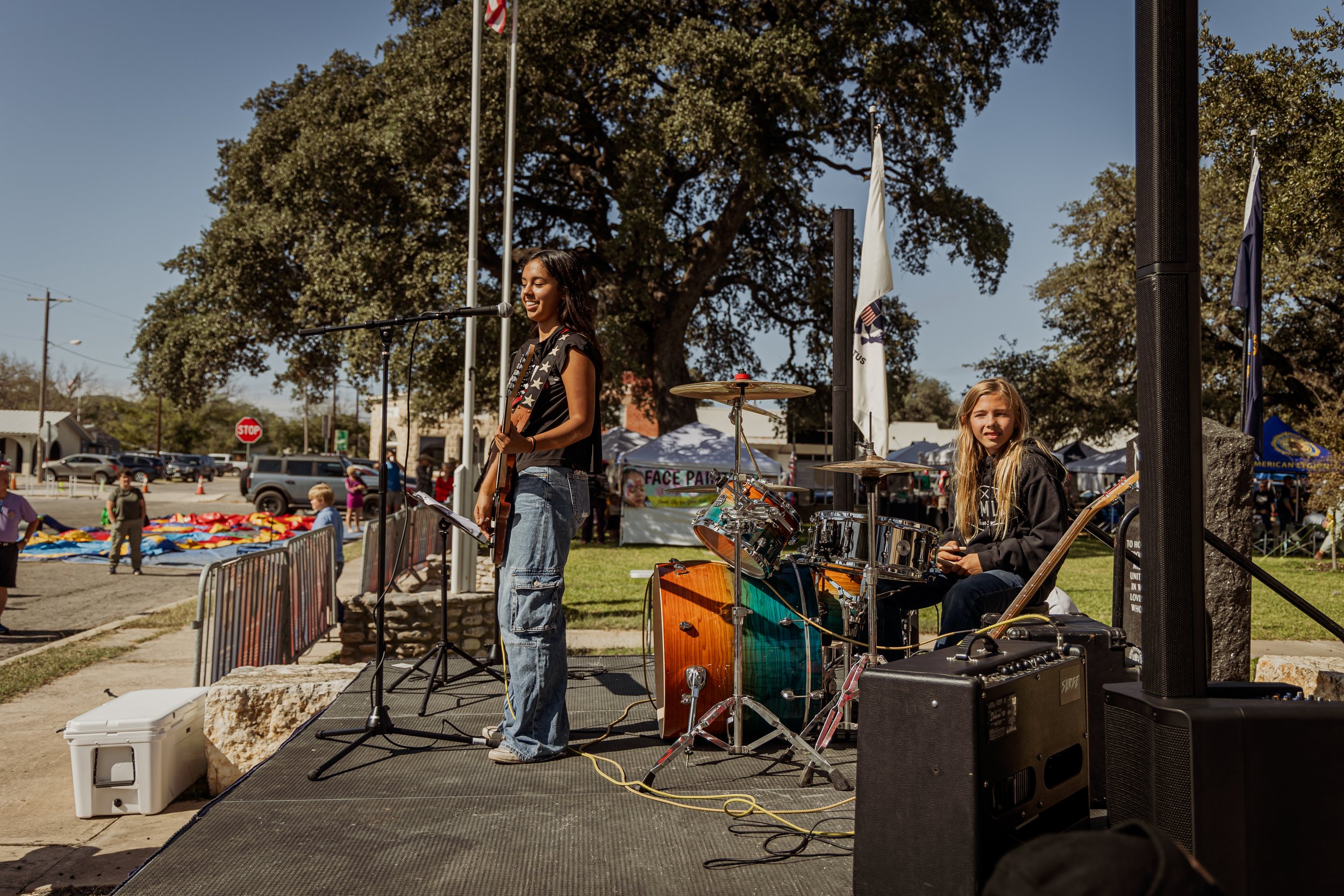 A young girl playing drums and a woman singing into a microphone while performing at an outdoor event on a sunny day.