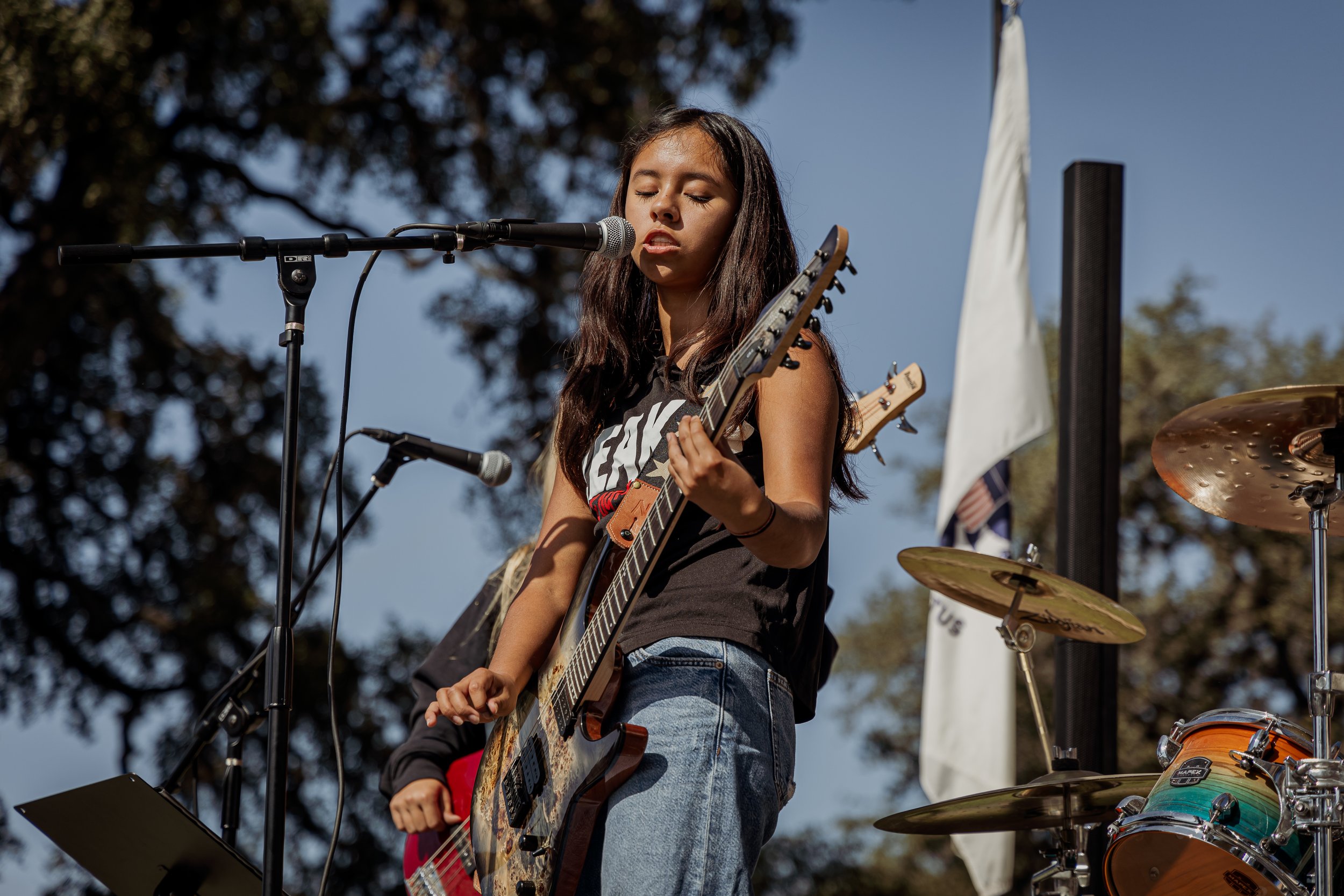 A young woman with long dark hair sings into a microphone while playing an electric guitar, standing outdoors during daytime with trees in the background.