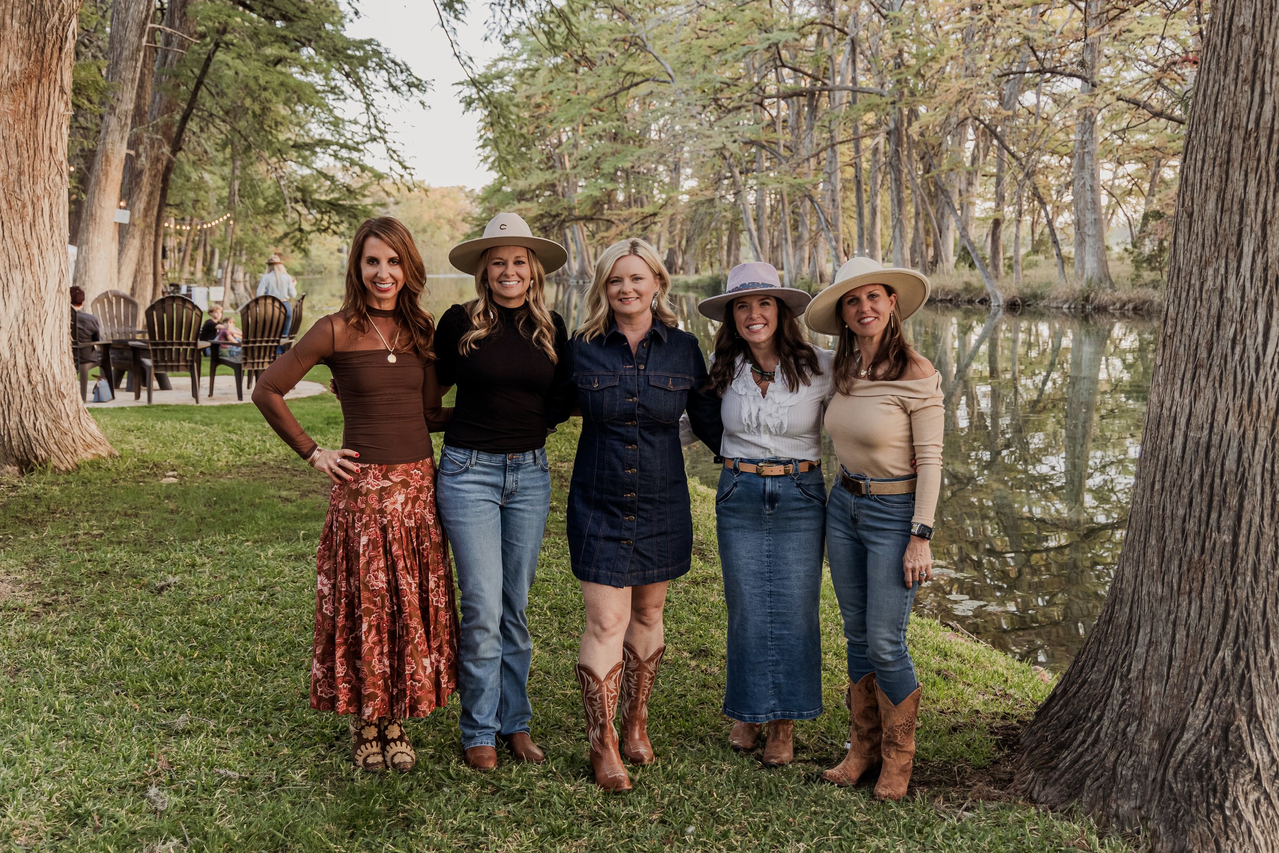 Five women standing outdoors near a lake with trees, dressed in casual country or ranch style with hats, jeans, and boots.