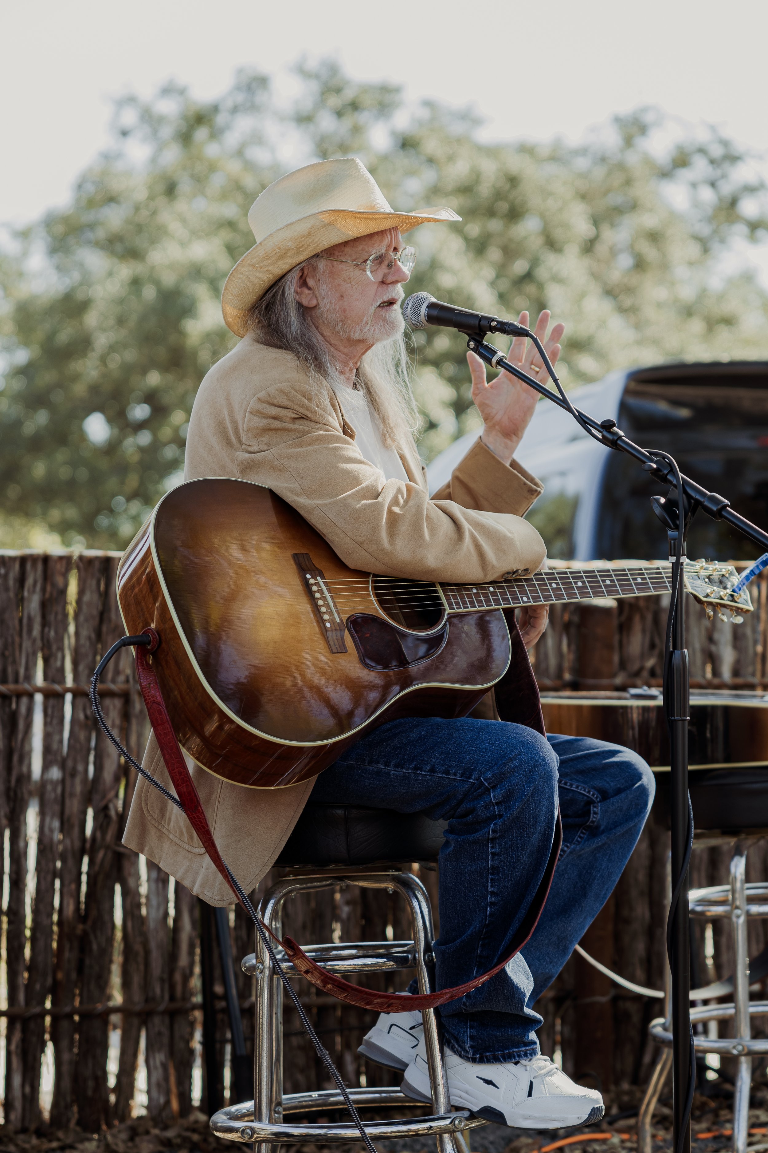 An older man with long gray hair and glasses, wearing a cowboy hat, beige jacket, and jeans, is sitting on a stool outdoors, playing an acoustic guitar and singing into a microphone.