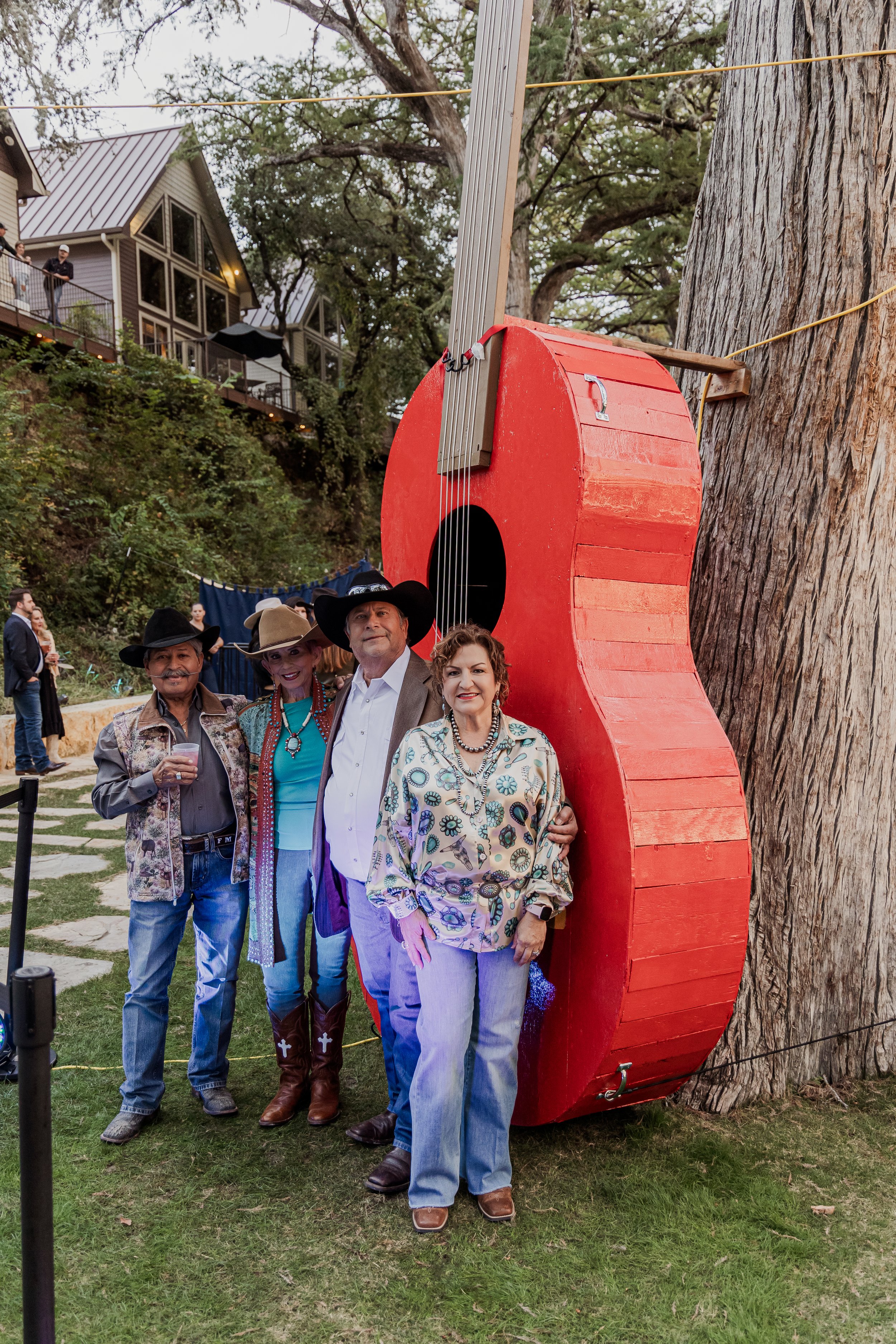 Four people dressed in cowboy and western-themed clothing standing under a large, red, guitar-shaped sculpture attached to a tree.