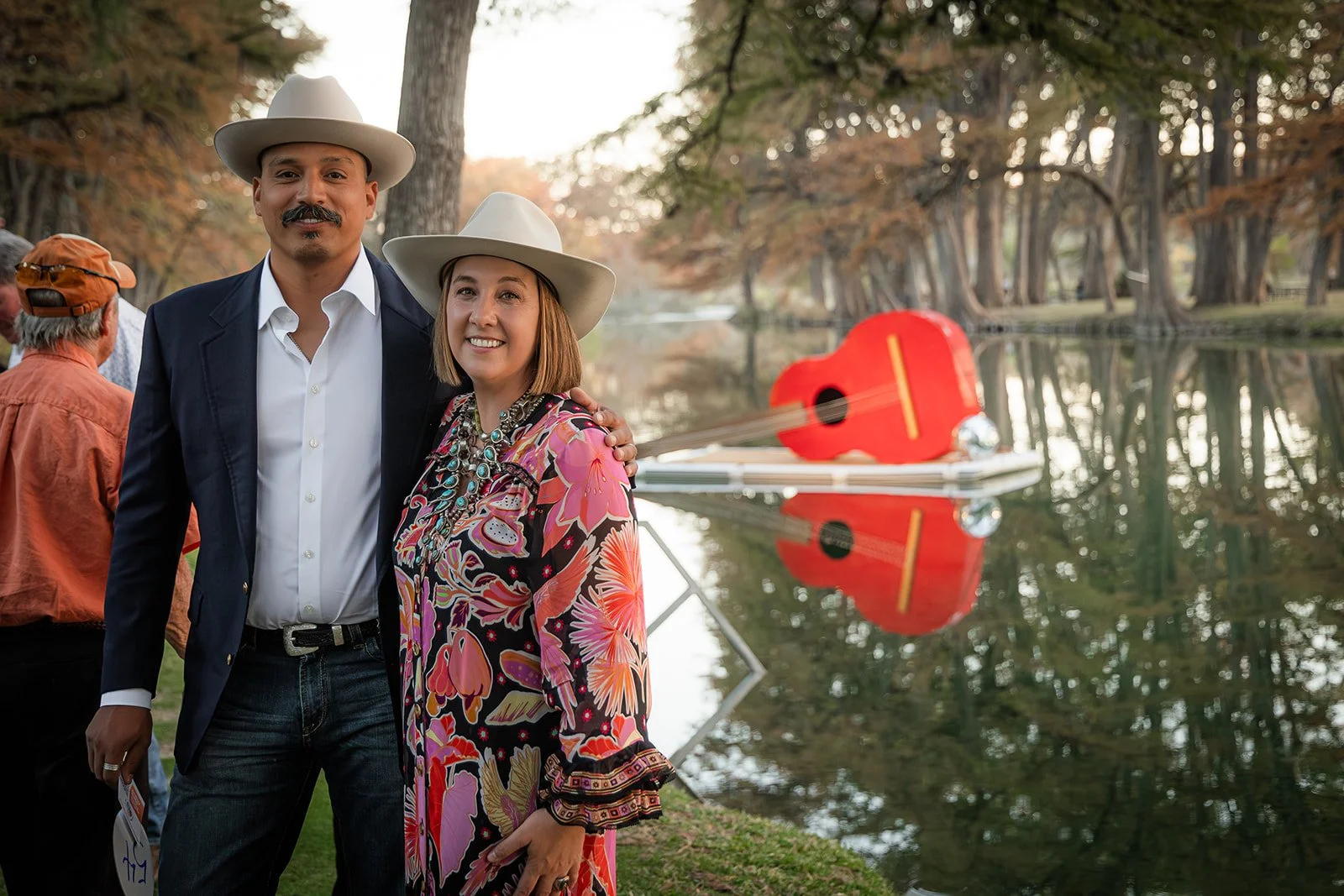 A smiling man and woman, both wearing white cowboy hats, standing by a lake during fall. The woman is wearing a colorful floral dress with statement jewelry, and the man is in a dark blazer with a white shirt. There are other people in the background