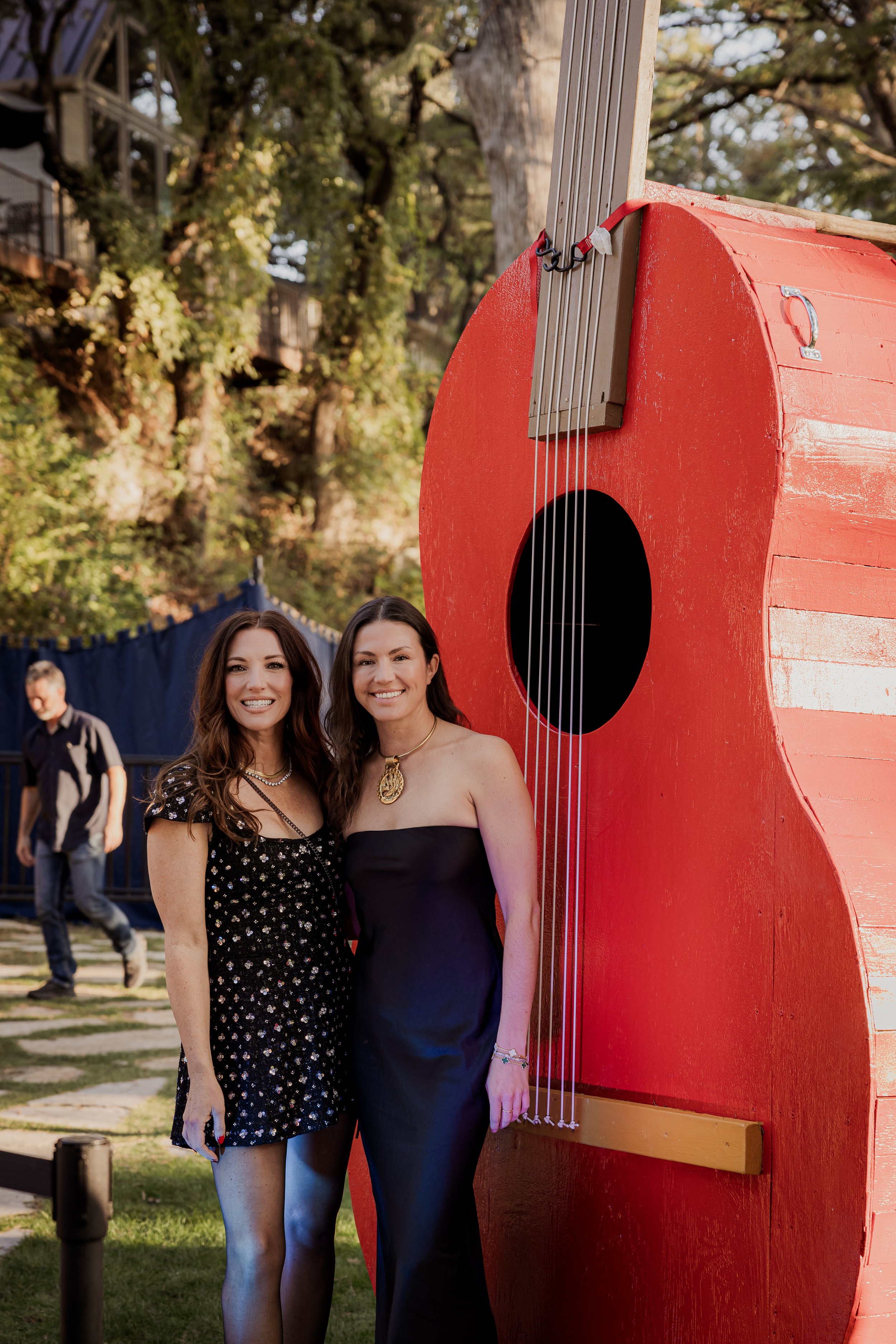 Two women standing outdoors near a large red guitar-shaped structure, smiling at the camera, with a man walking in the background and trees surrounding the area.