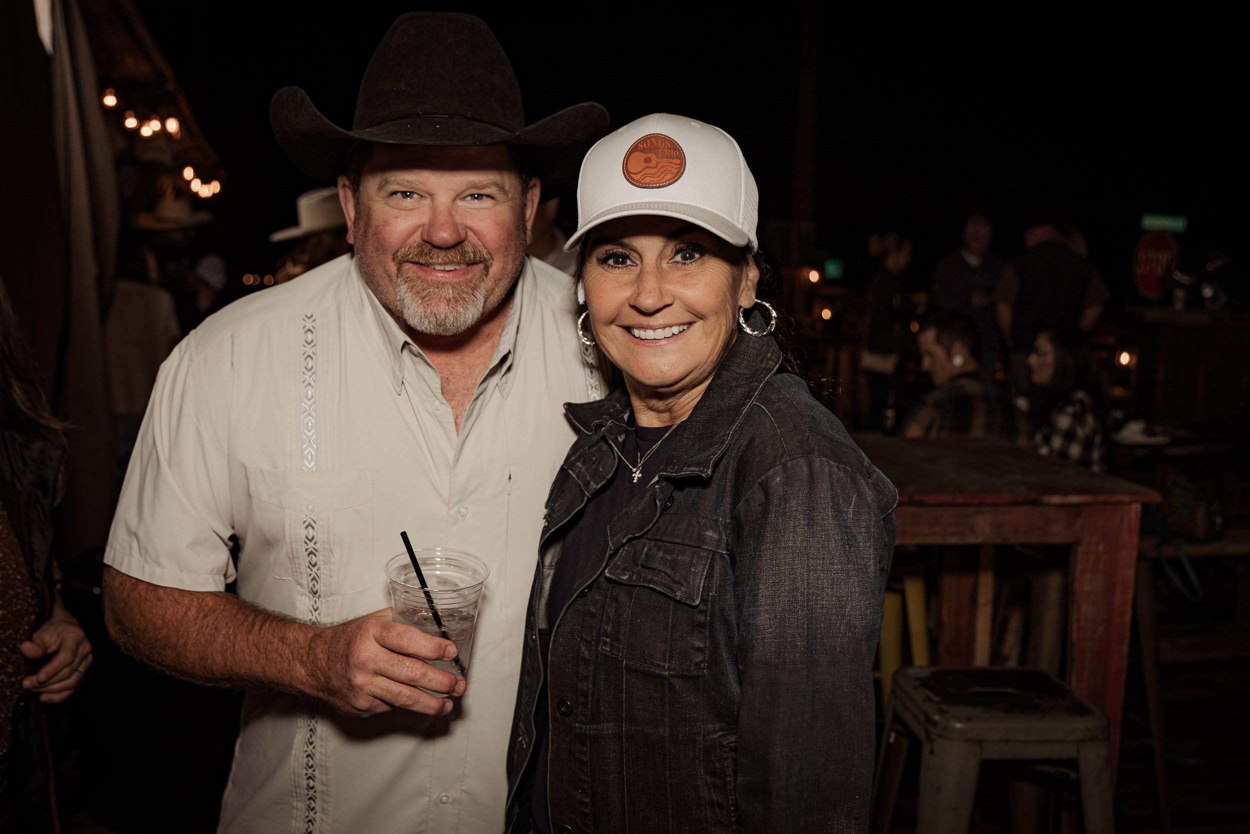 A smiling man wearing a cowboy hat and a white shirt standing next to a smiling woman wearing a baseball cap, hoop earrings, and a dark denim jacket at a nighttime outdoor event.