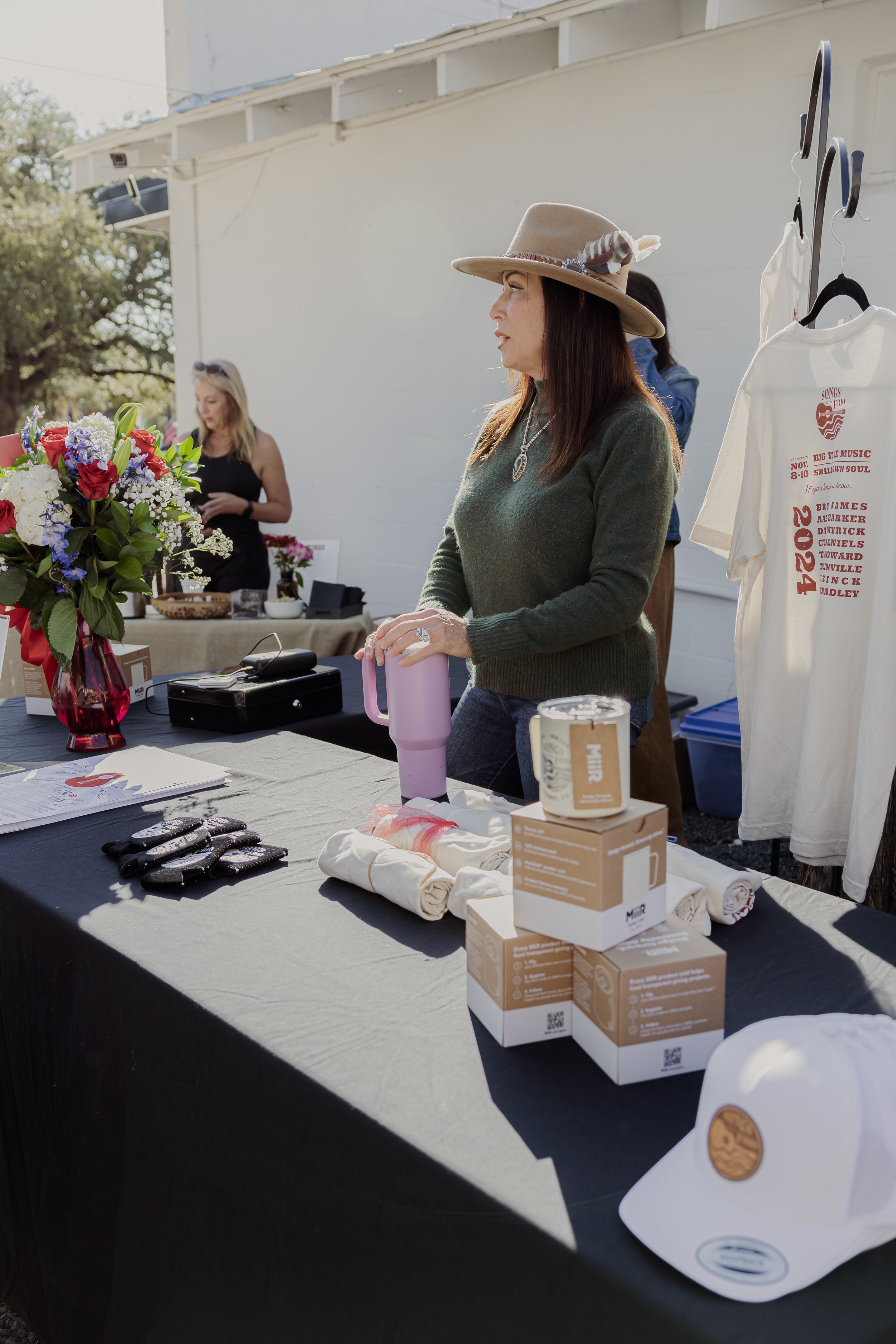 Woman in a wide-brimmed hat at a vendor stand with various items including a bouquet of flowers, boxed mugs, rolled textiles, and a hat. Two other women are in the background at the outdoor event.