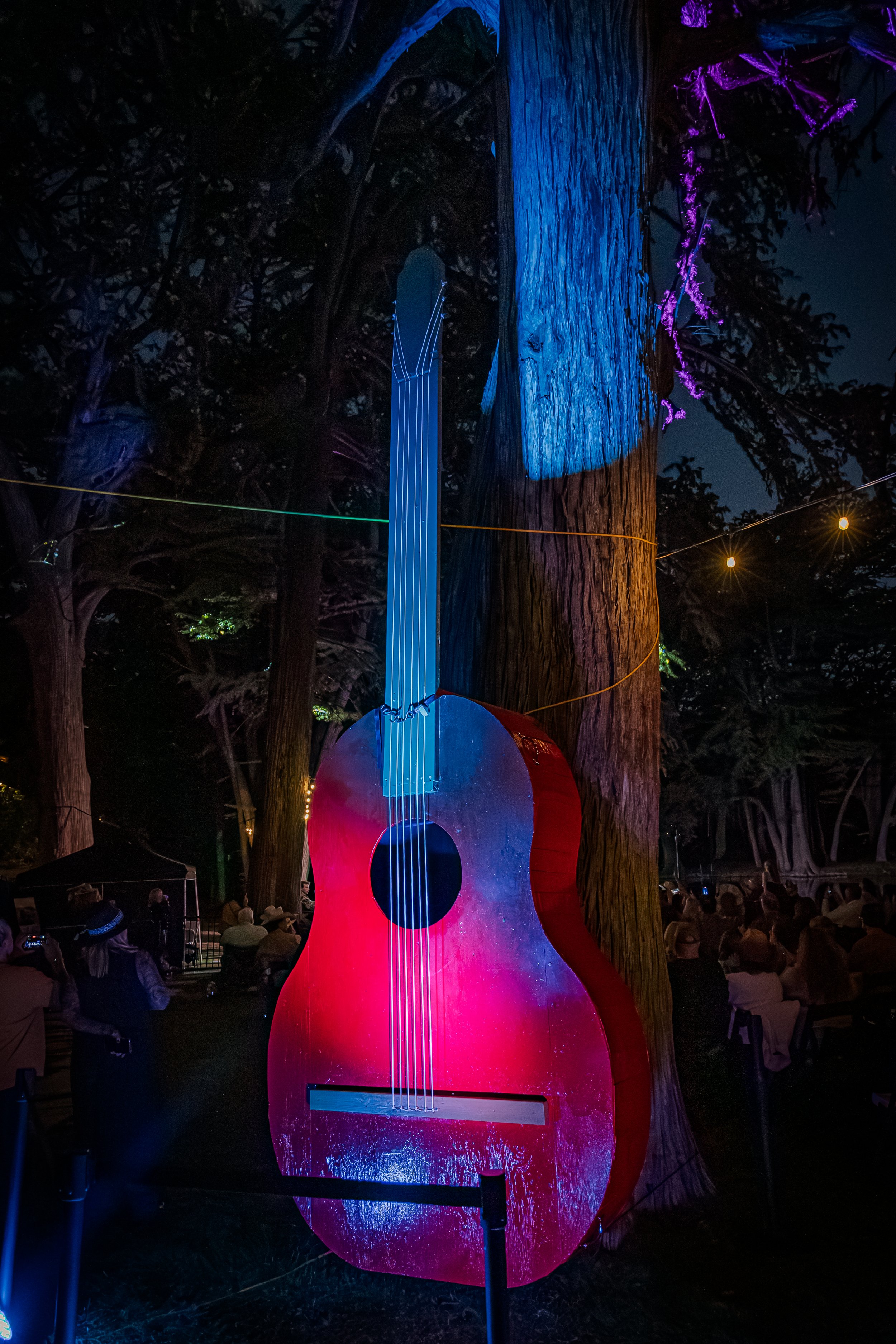 A large, illuminated guitar sculpture displayed outdoors at night, tied to a tree, with people seated in the background.