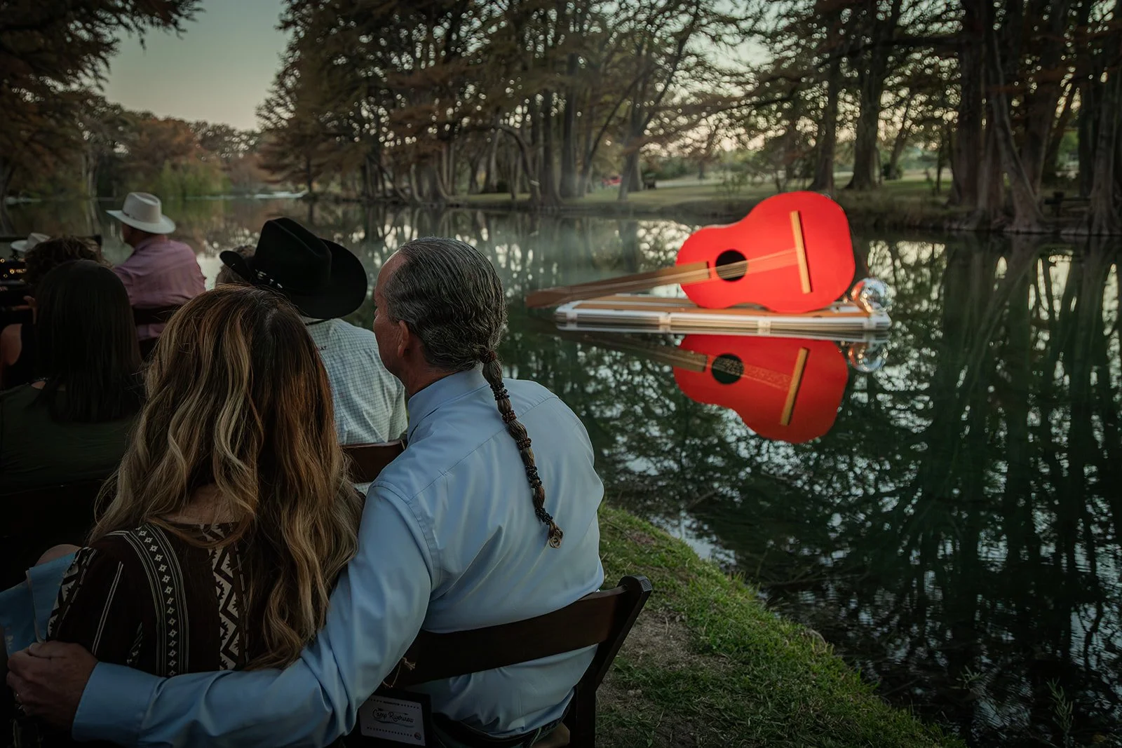 People sitting outdoors near a lake at sunset, with a large red guitar floating on a floating platform in the water and surrounded by trees.