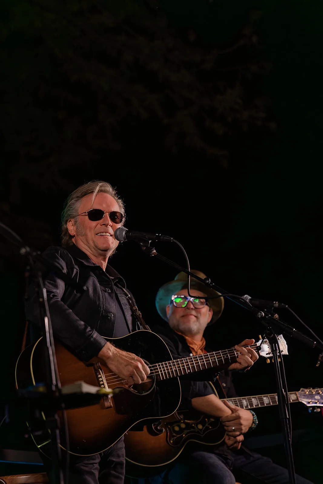 Two musicians performing on stage, one is singing into a microphone and playing an acoustic guitar, the other is playing an electric guitar and wearing glasses and a hat.