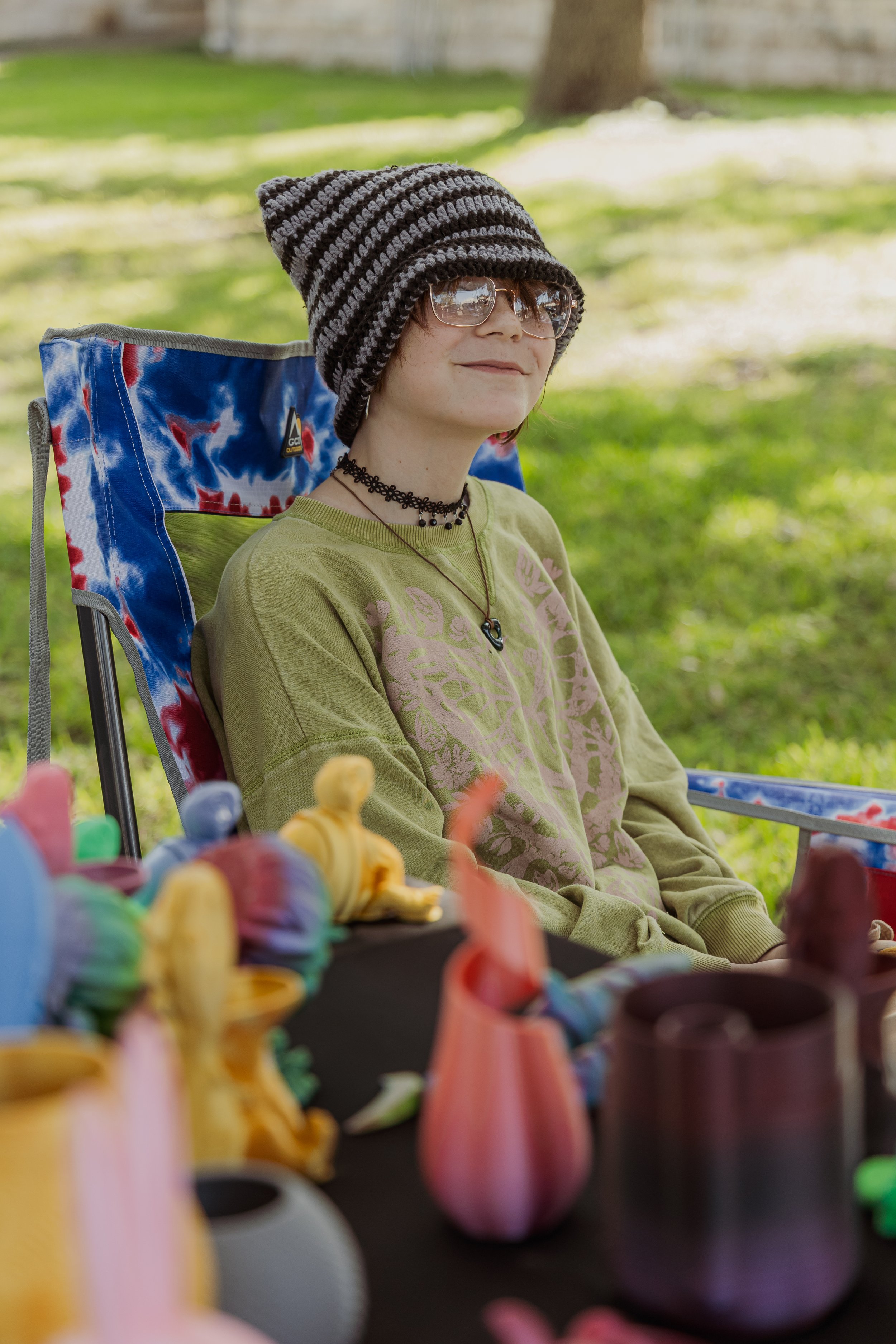 Young woman with glasses, striped knit hat, and layered necklaces sitting outdoors in a lawn chair at a table with colorful art sculptures and vases.
