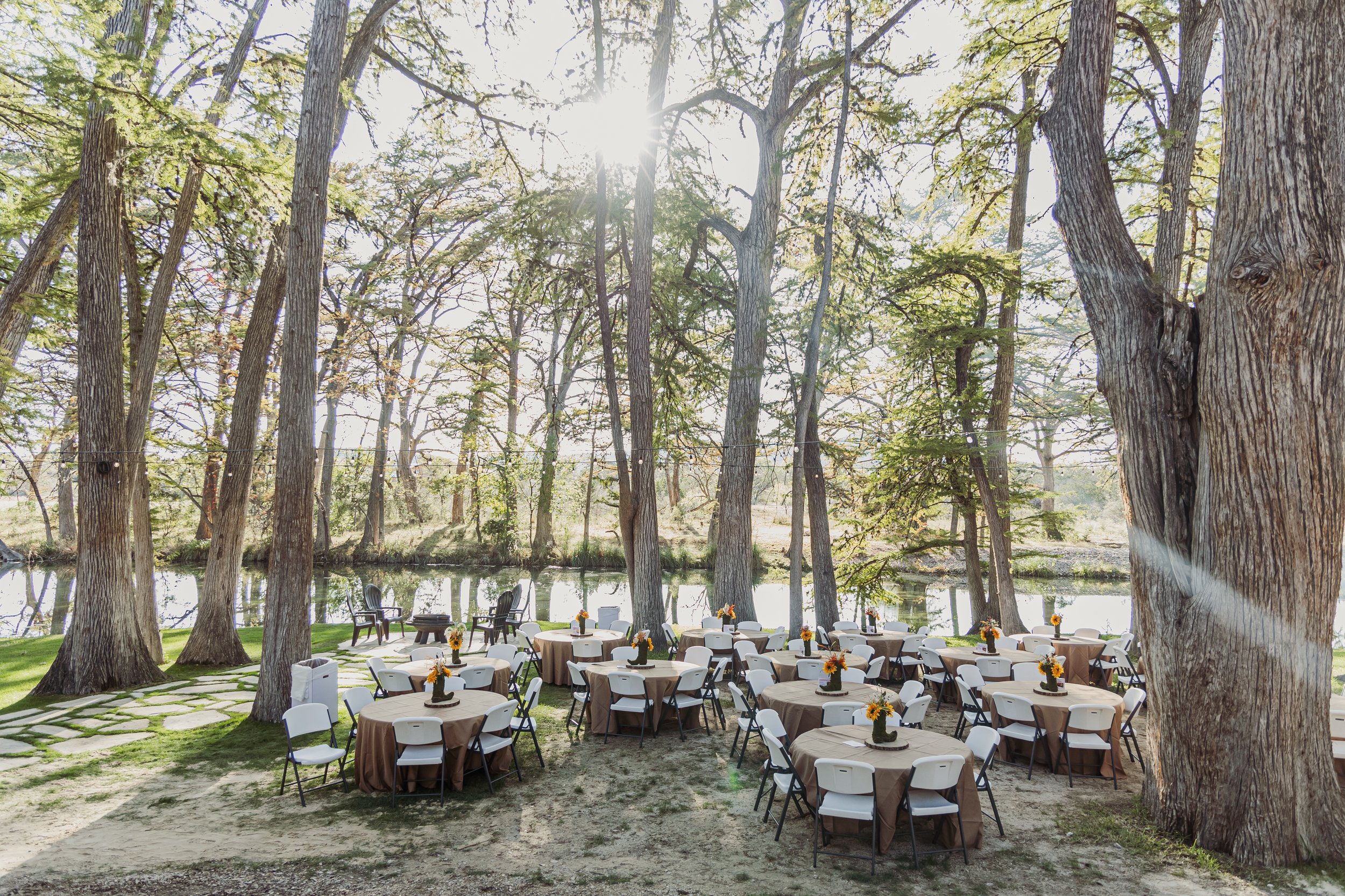 Outdoor event setup with round tables covered in brown tablecloths, decorated with small flower arrangements, surrounded by white and black chairs, set among large trees near a river with sunlight filtering through the leaves.