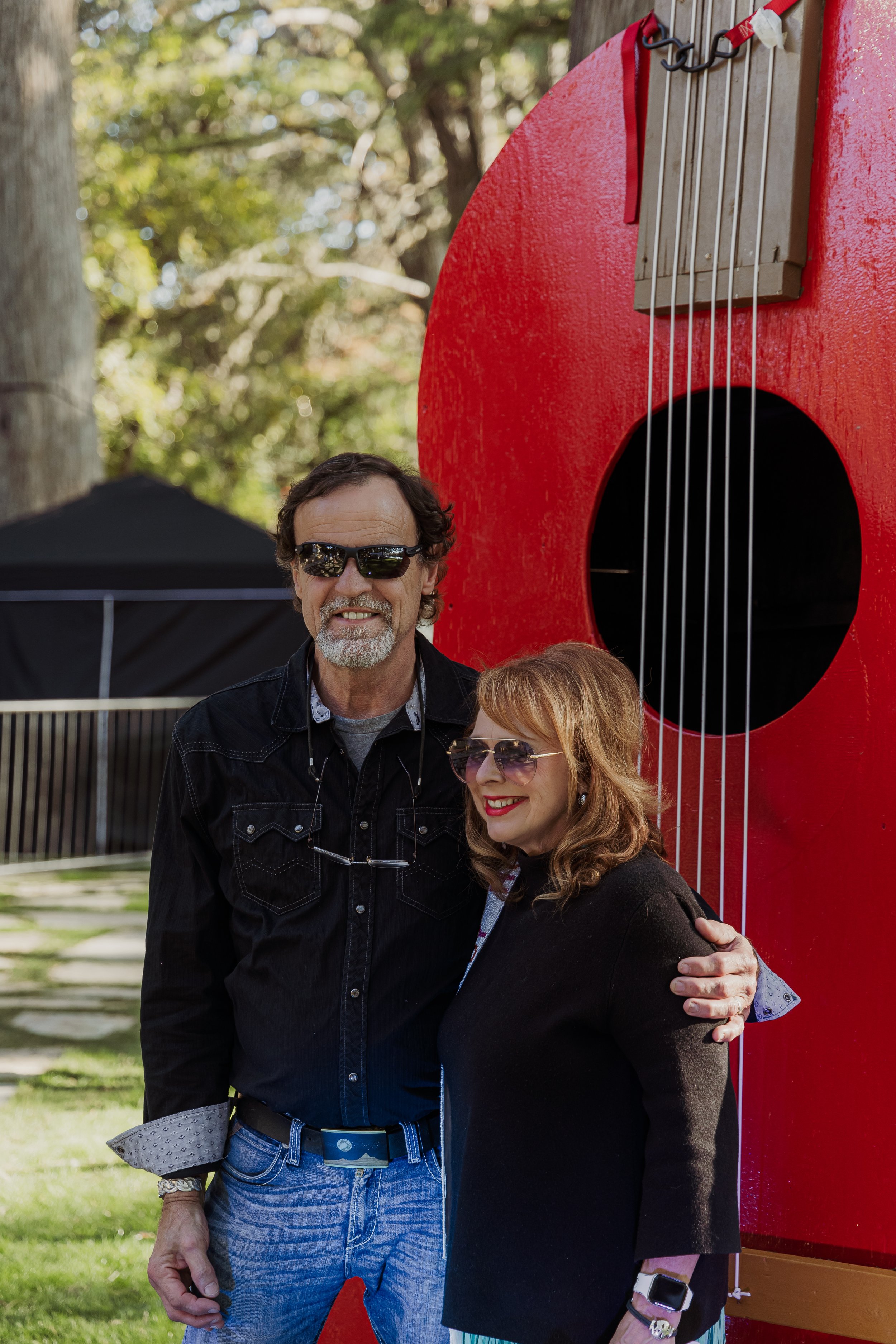 A man and woman wearing sunglasses standing outside on grass, smiling, with a large red guitar sculpture behind them and trees in the background.