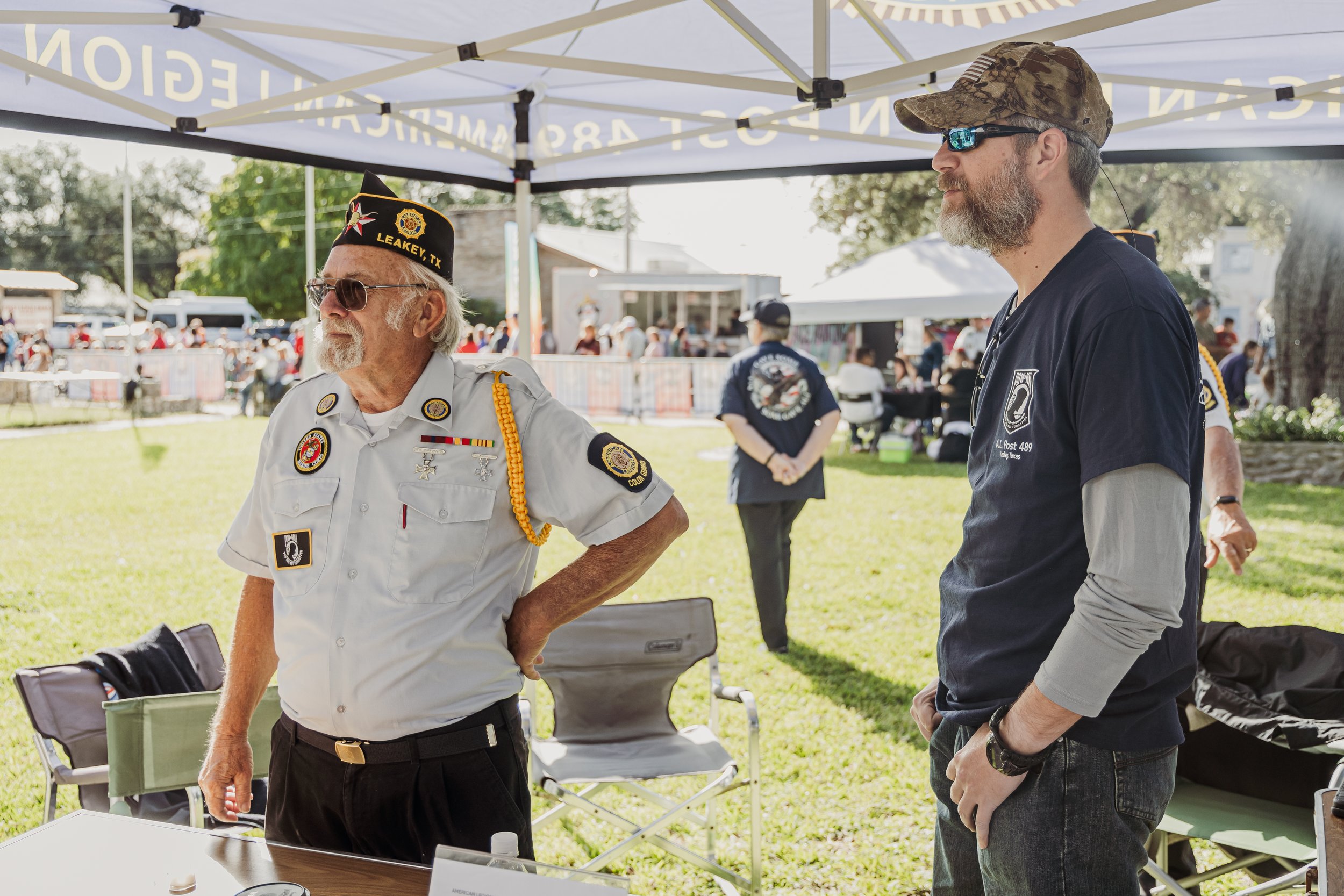 Two men talking at an outdoor event under a white canopy tent, with other people and tents visible in the background.