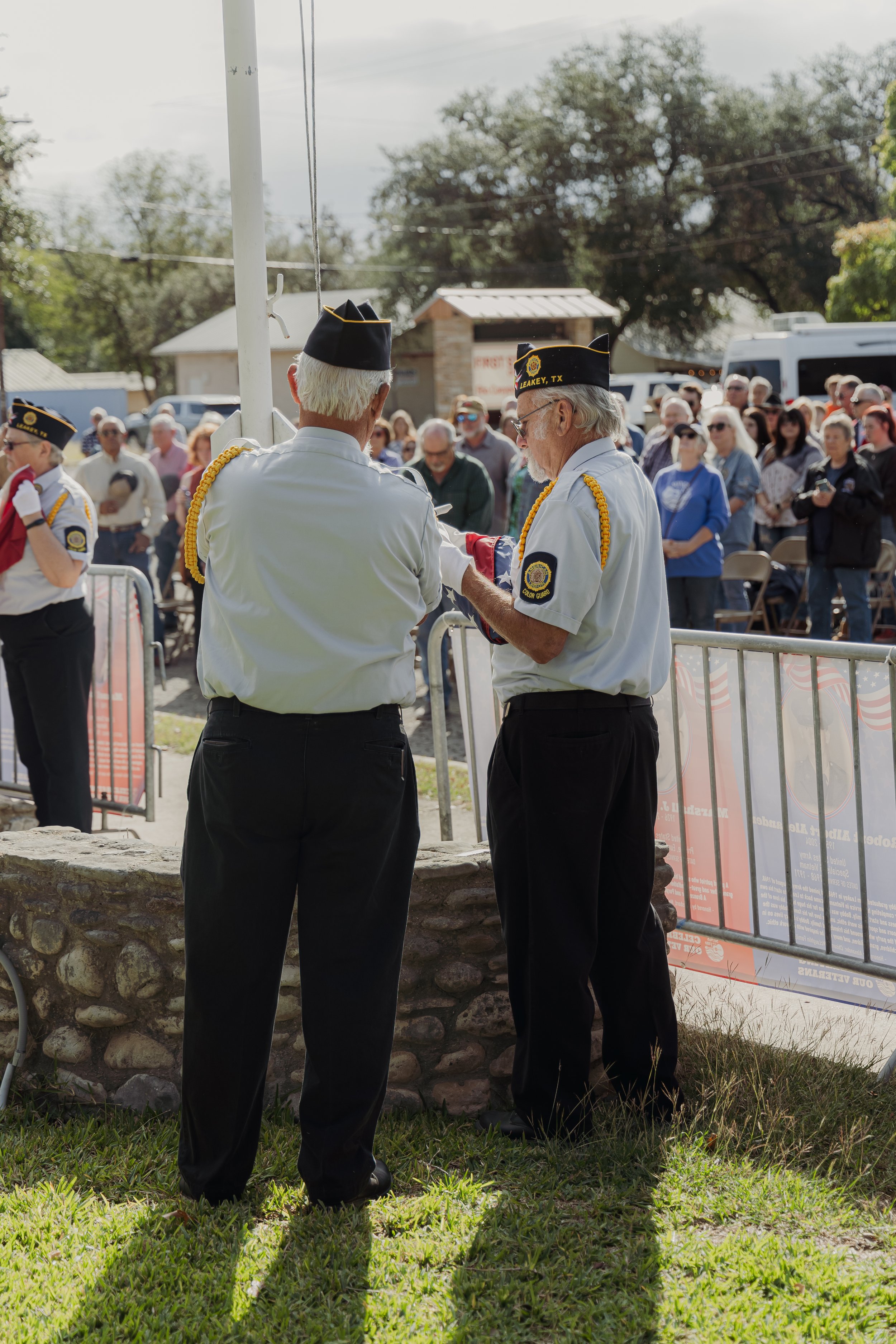 Two elderly men in the U.S. flag honor guard uniform, standing outdoors in front of a crowd at a Memorial Day or Veterans Day event. One man is wearing a black cap with yellow text, and both have yellow cords on their shirts. They are reading from a