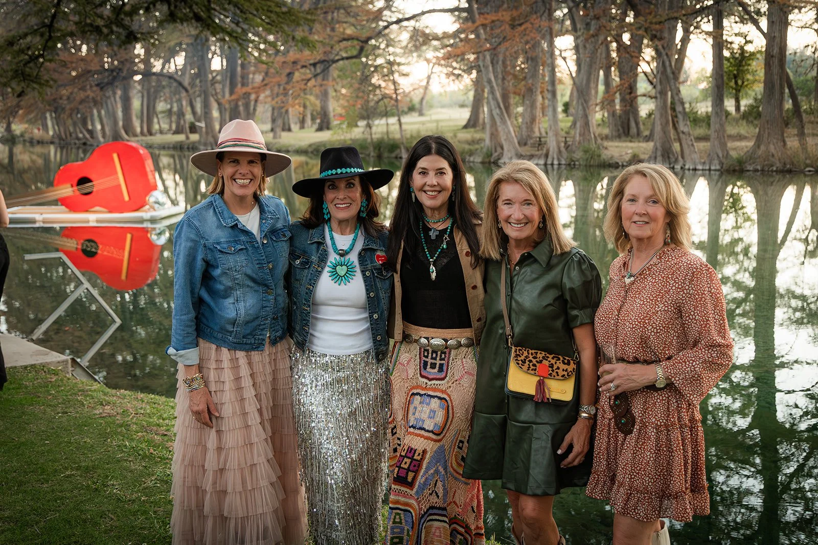 Five women standing together outdoors near a lake, smiling, with trees and a sunset in the background. One woman is holding a guitar, and there is a red guitar-shaped boat on the water.