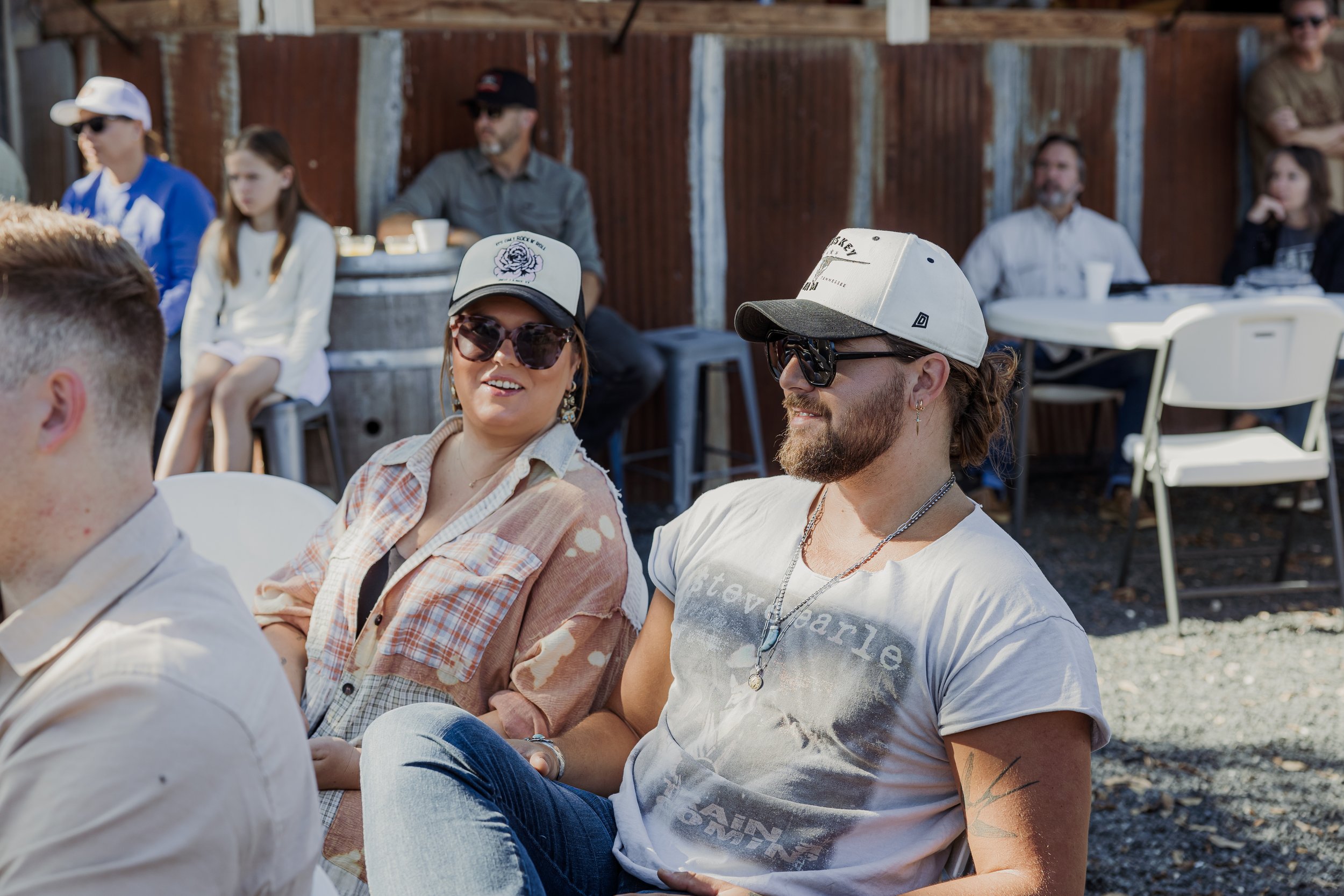 People sitting outdoors at a gathering or event, some wearing sunglasses and hats, in front of a wooden fence.
